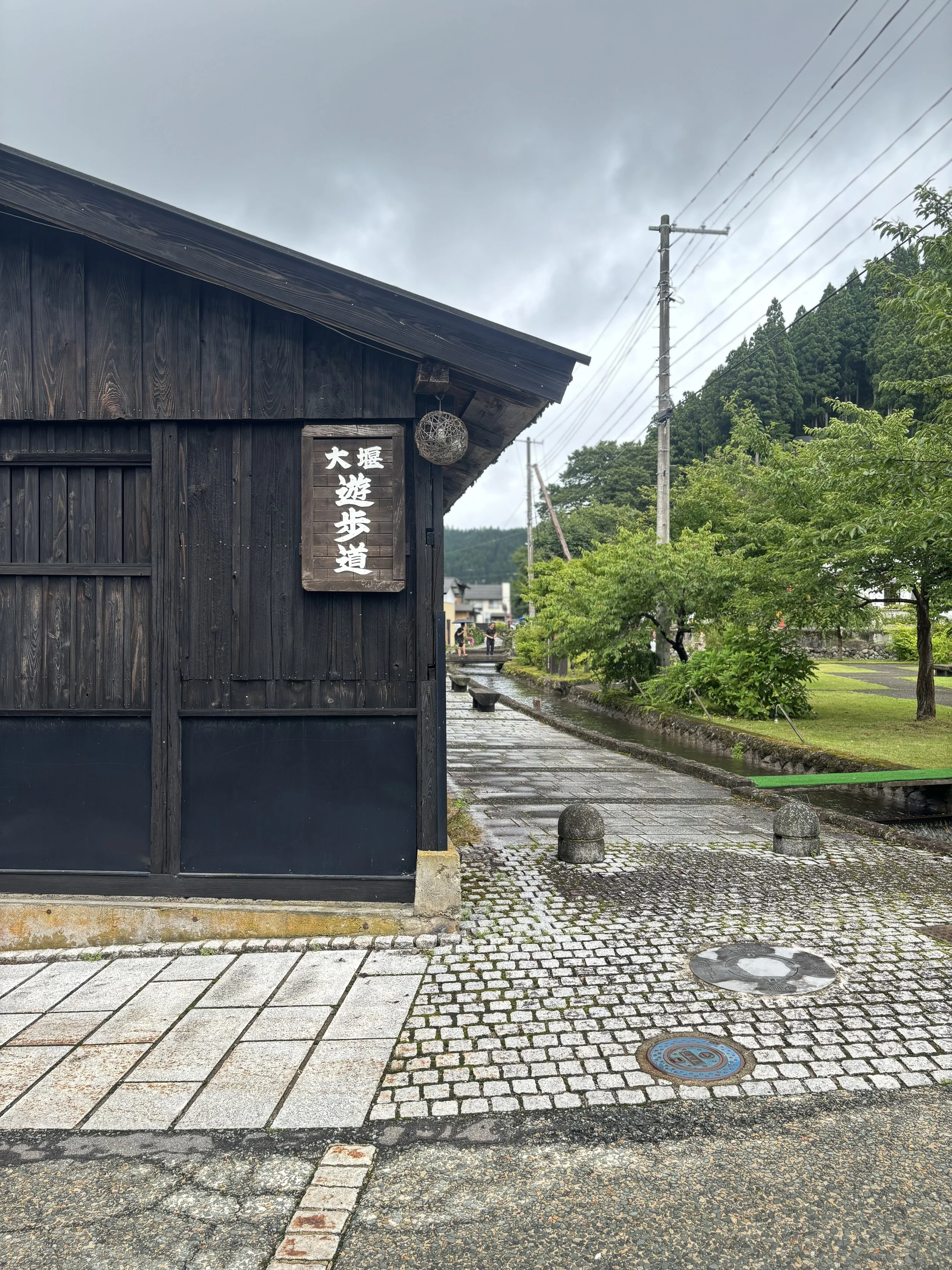 A street scene featuring a traditional black wooden building with Japanese characters on a sign, surrounded by greenery and power lines under a cloudy sky.
