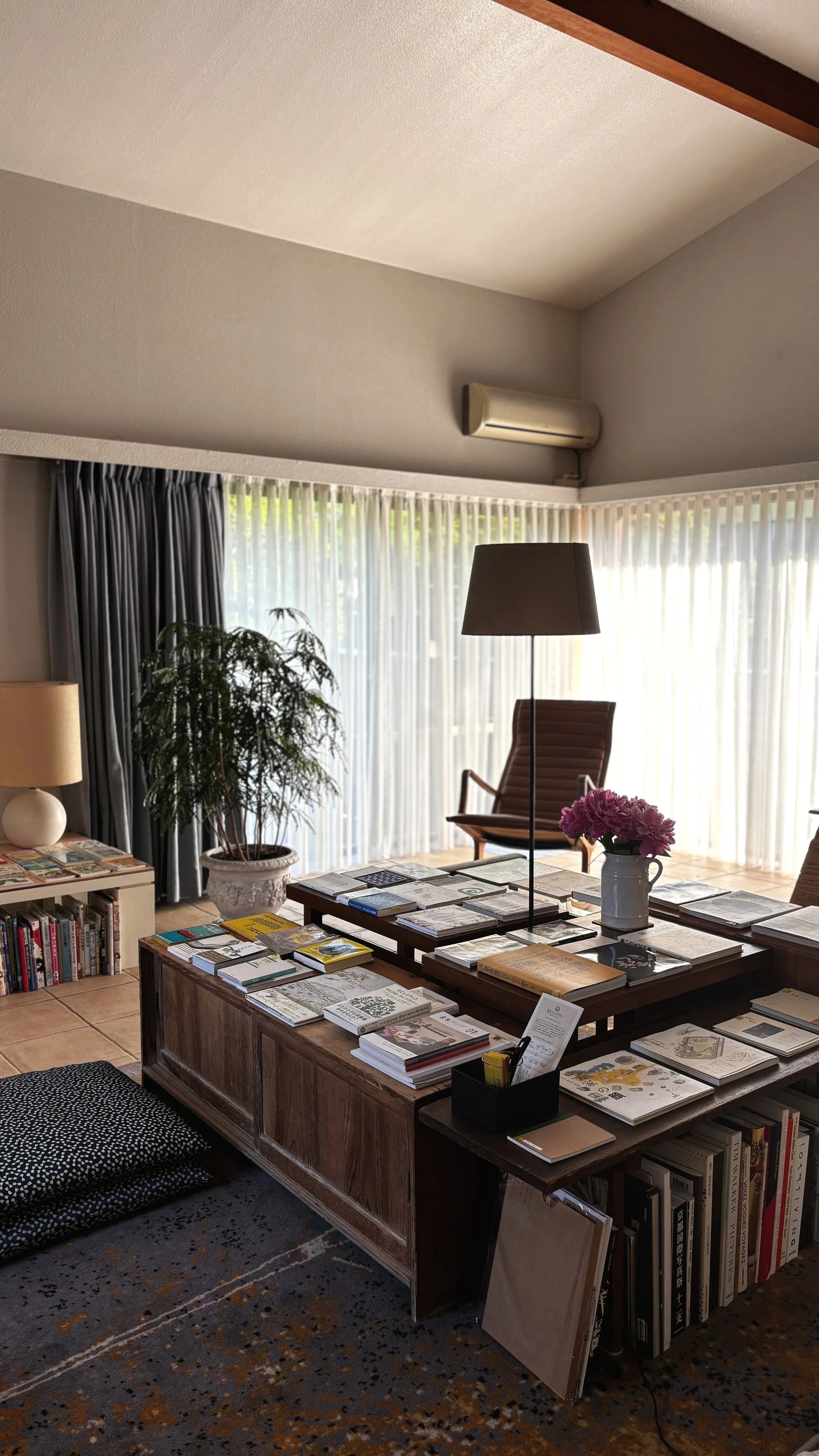 A cozy living room with large windows with sheer curtains, a wooden coffee table filled with books and magazines, a potted plant, a side table with a lamp, and a tall floor lamp, with a comfortable armchair and a reading chair.