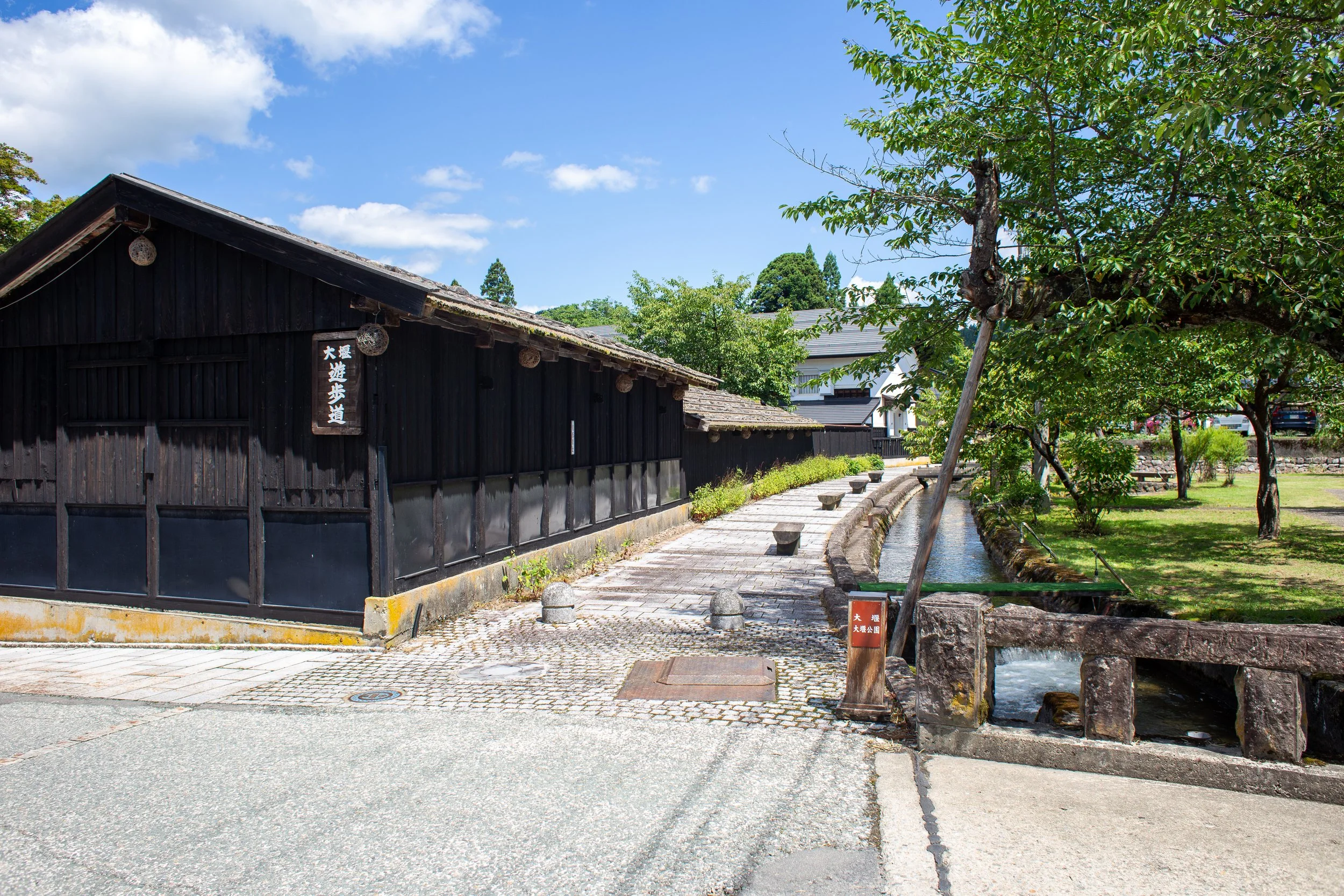 A traditional Japanese black wooden building with a paved pathway alongside a small stream, surrounded by green trees and clear blue sky.
