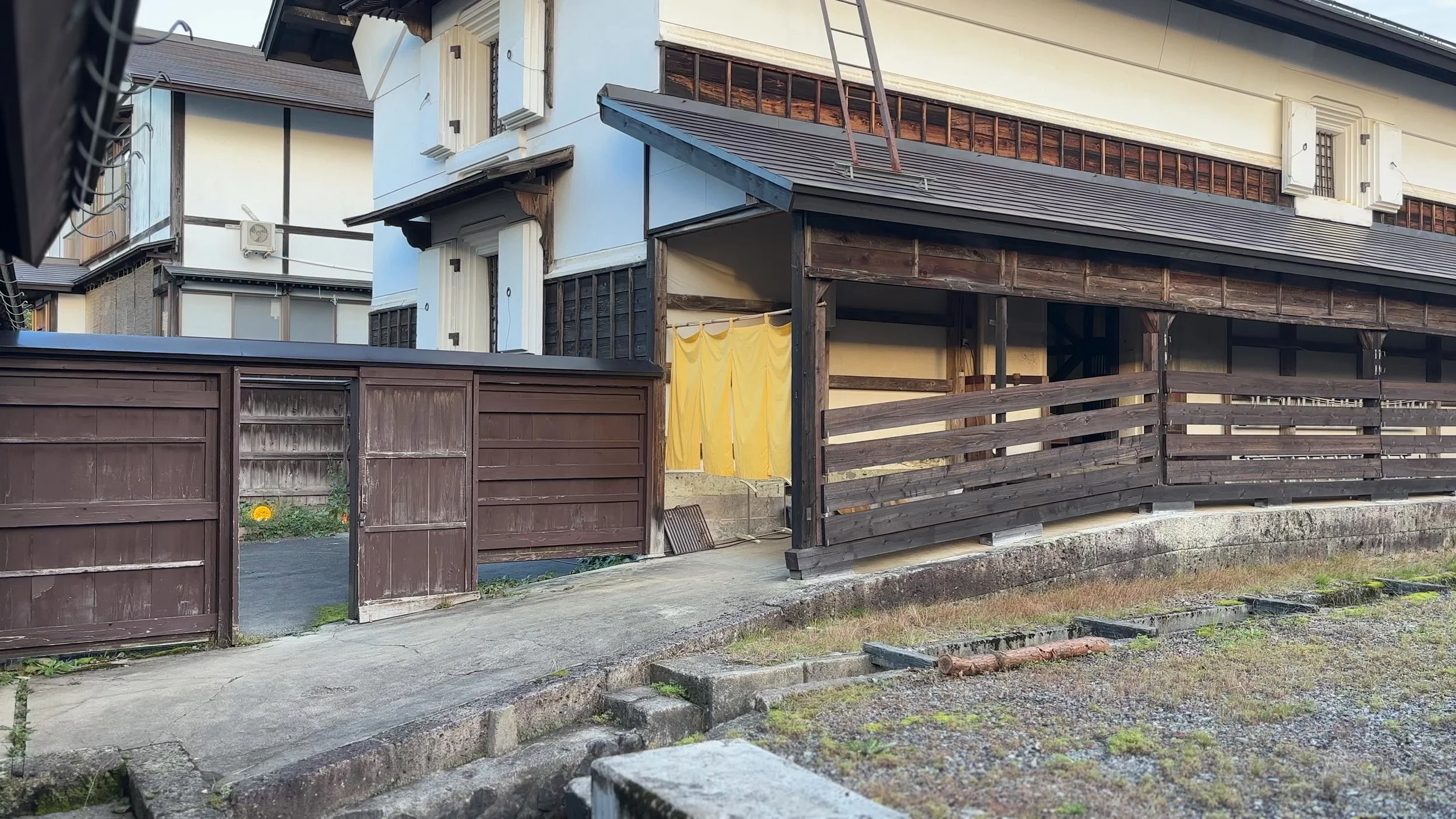 Traditional Japanese house with wooden fence, yellow cloth hanging, and exterior stairs, in a residential area.