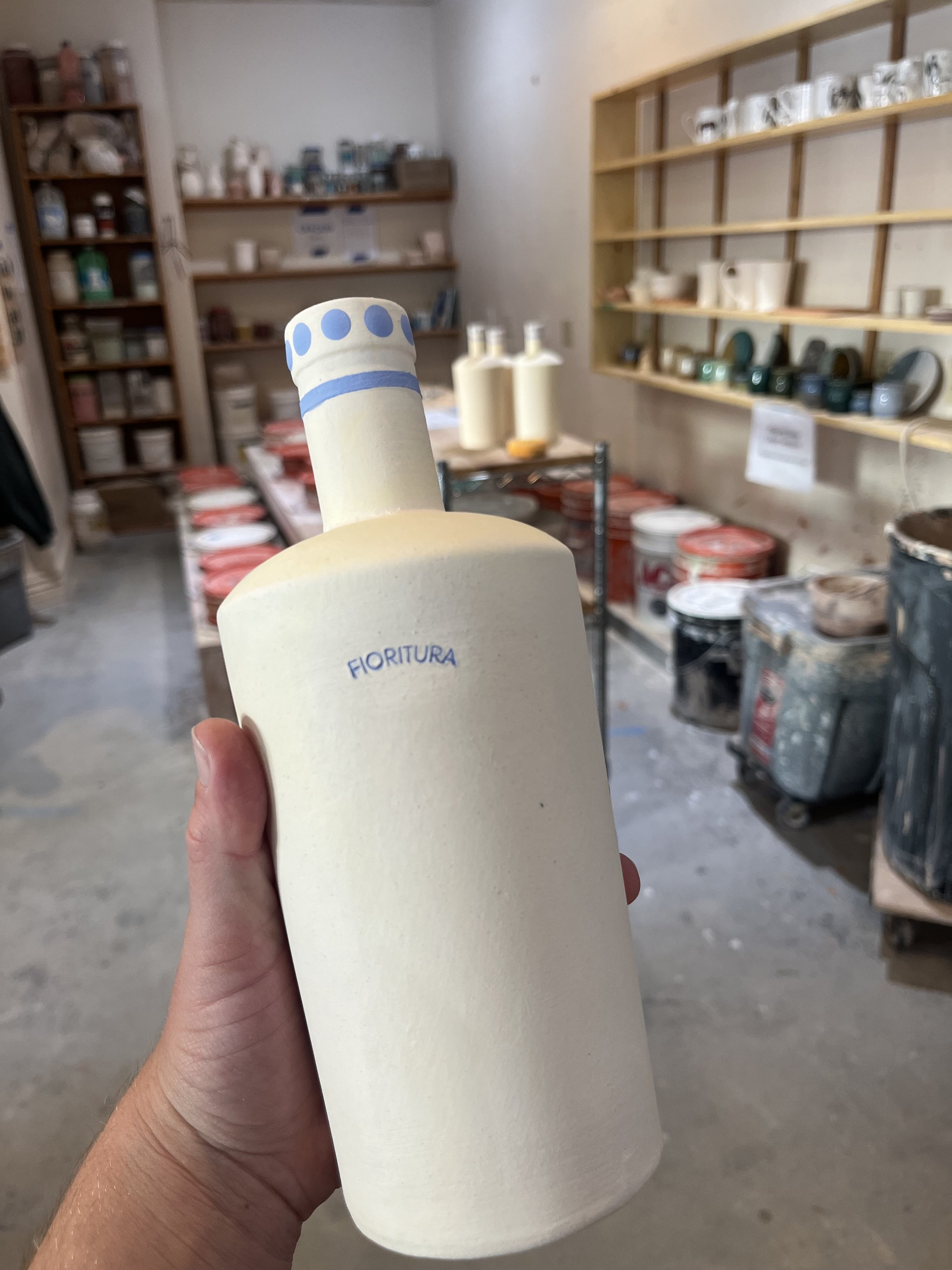 Person holding a ceramic bottle in a pottery workshop, with shelves and art supplies in the background.