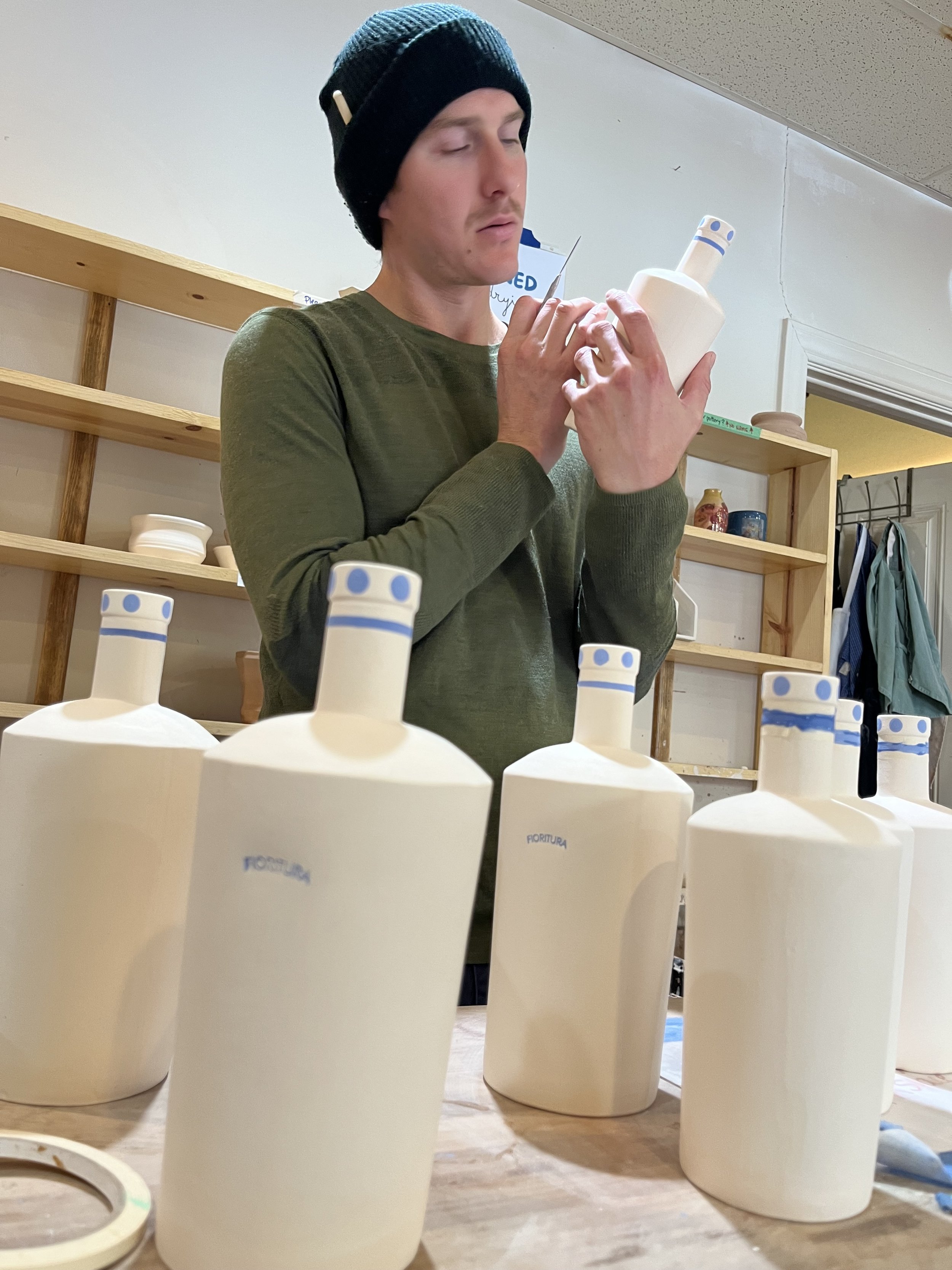 Person inspecting a ceramic bottle, wearing a green shirt and black beanie, surrounded by white ceramic bottles with blue accents in a studio.