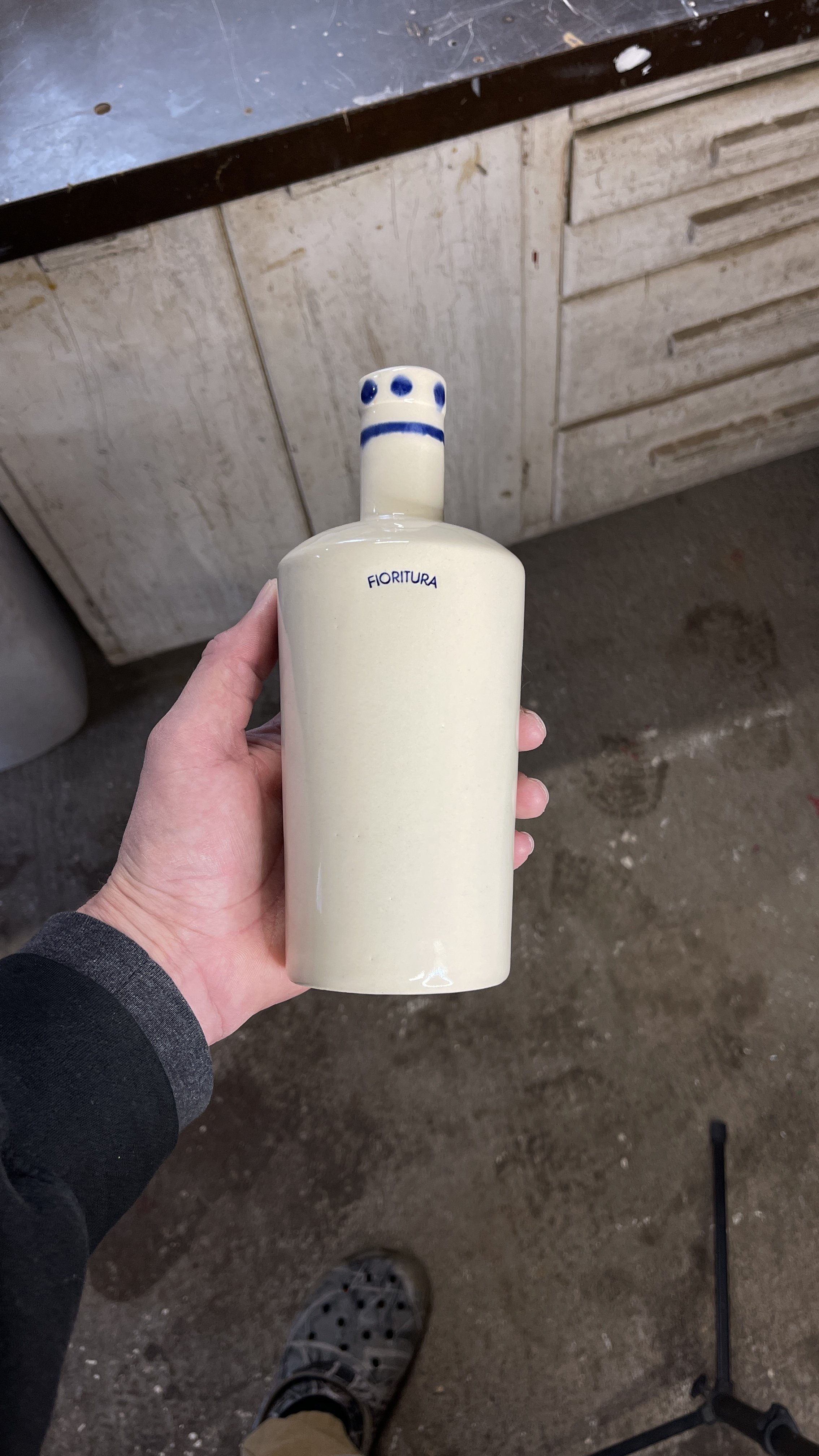 Person holding a beige ceramic bottle labeled "Fioritura" with a blue ring and dots near the top, against a workshop background.