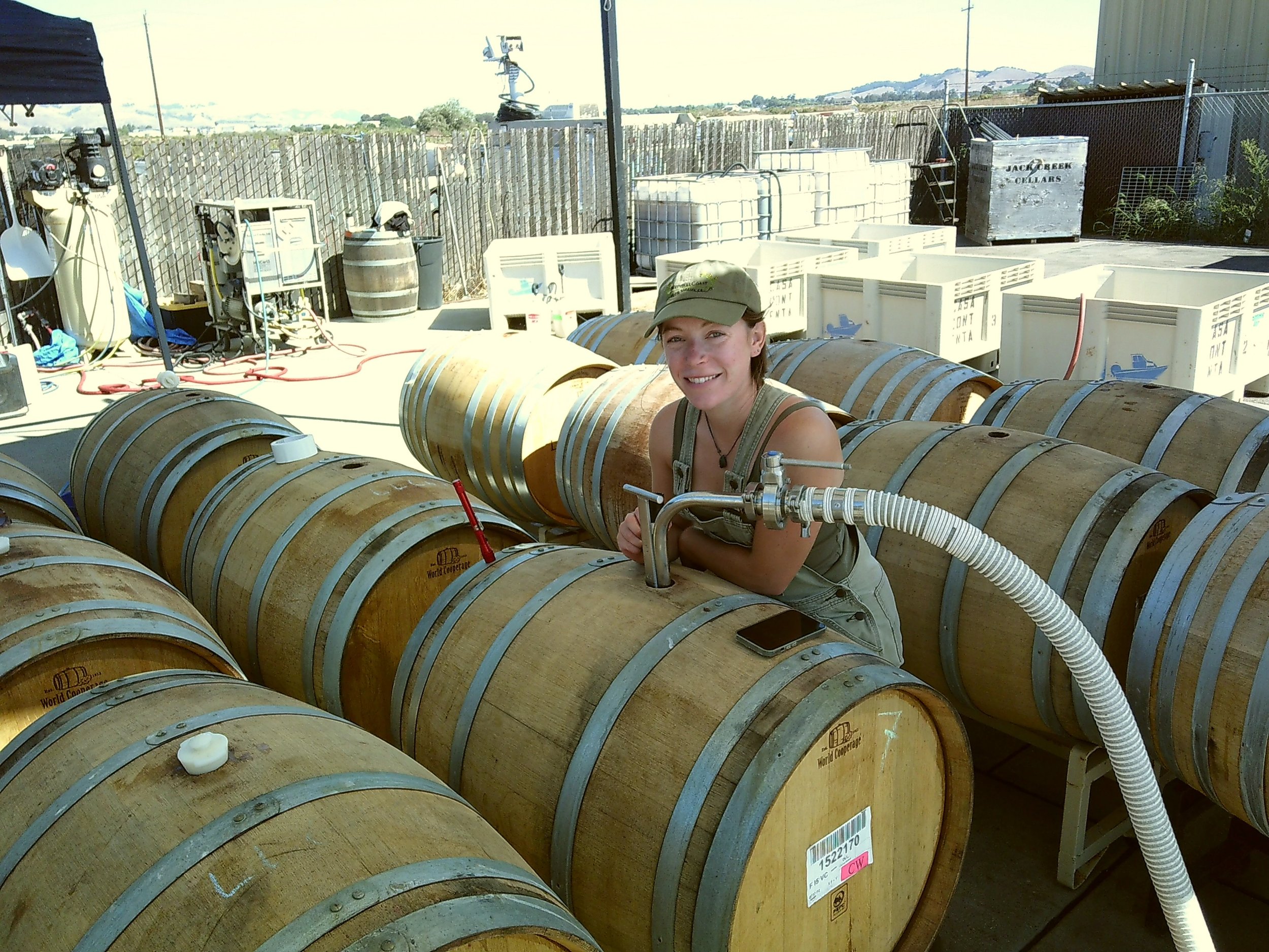 A person in a cap and overalls smiles while leaning on a stack of wooden barrels in a winery setting, with hoses and equipment nearby.