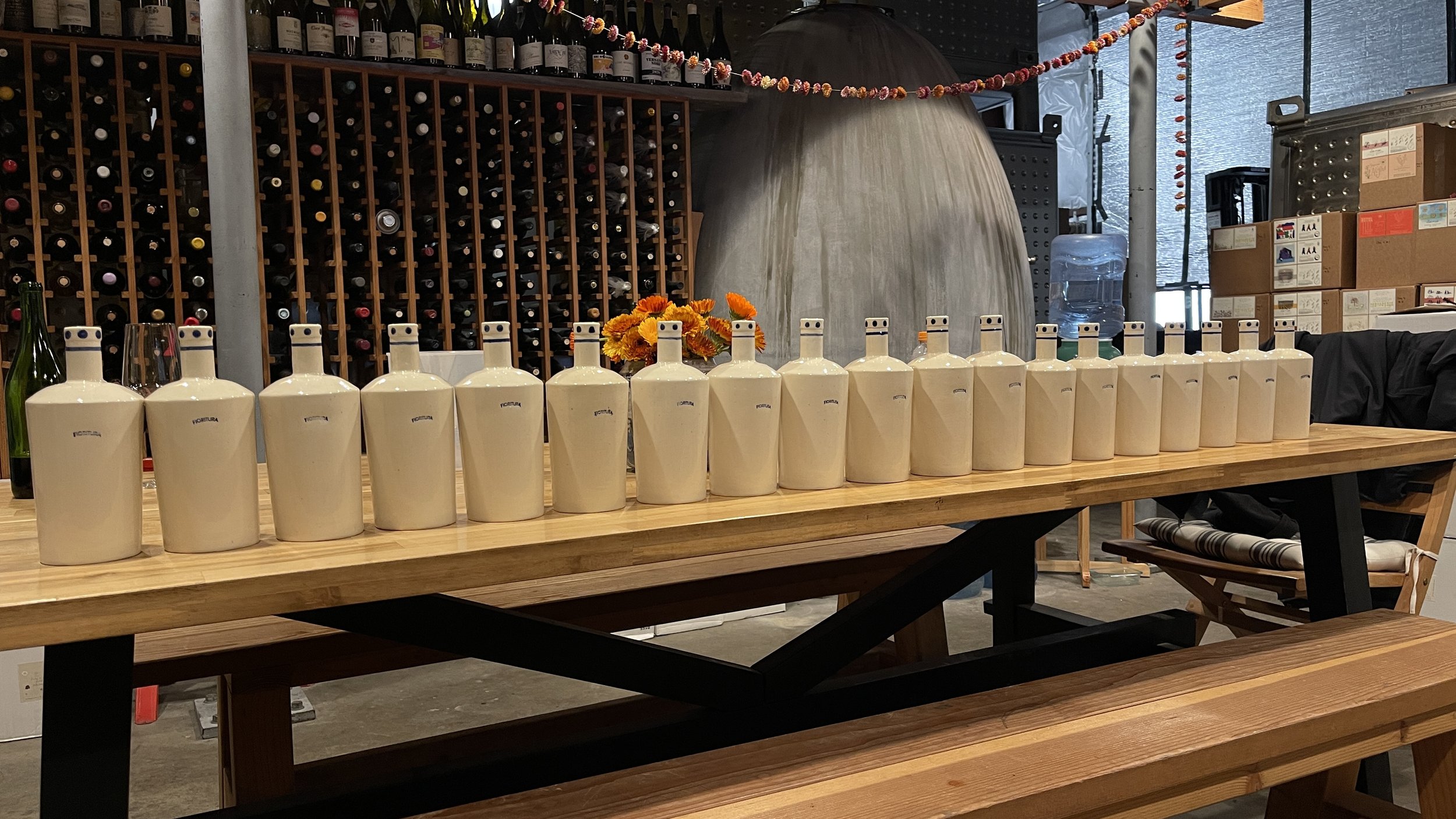 Row of identical ceramic bottles on a wooden table in a wine cellar, with wine racks and various bottles in the background.