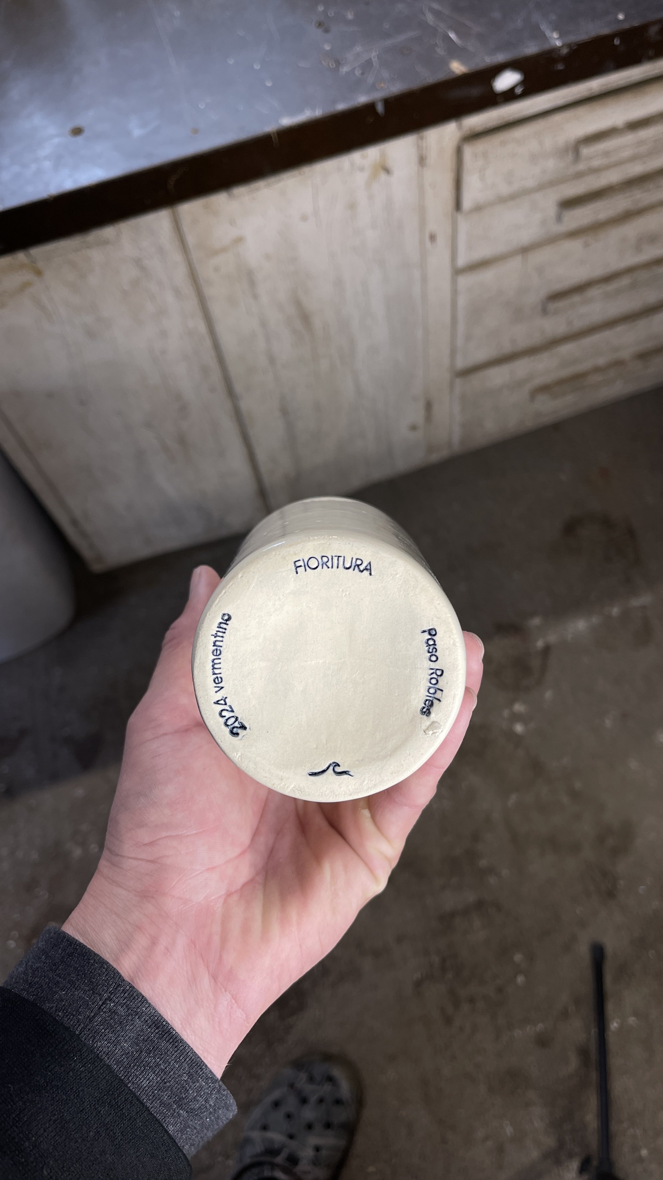 A person holding a ceramic cup with the words '2024 Vermentino', 'Fiortura', and 'Paso Robles' on the bottom. The background shows a workshop with a metal counter and wooden drawers.