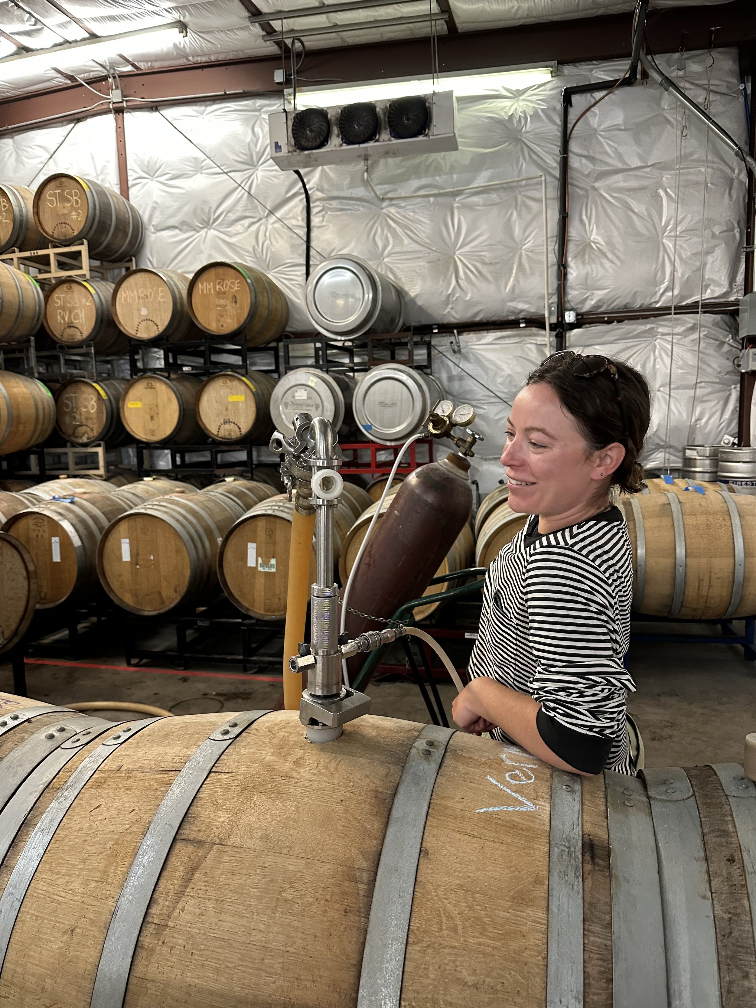 Person operating equipment on wine barrel in storage room with stacked barrels.