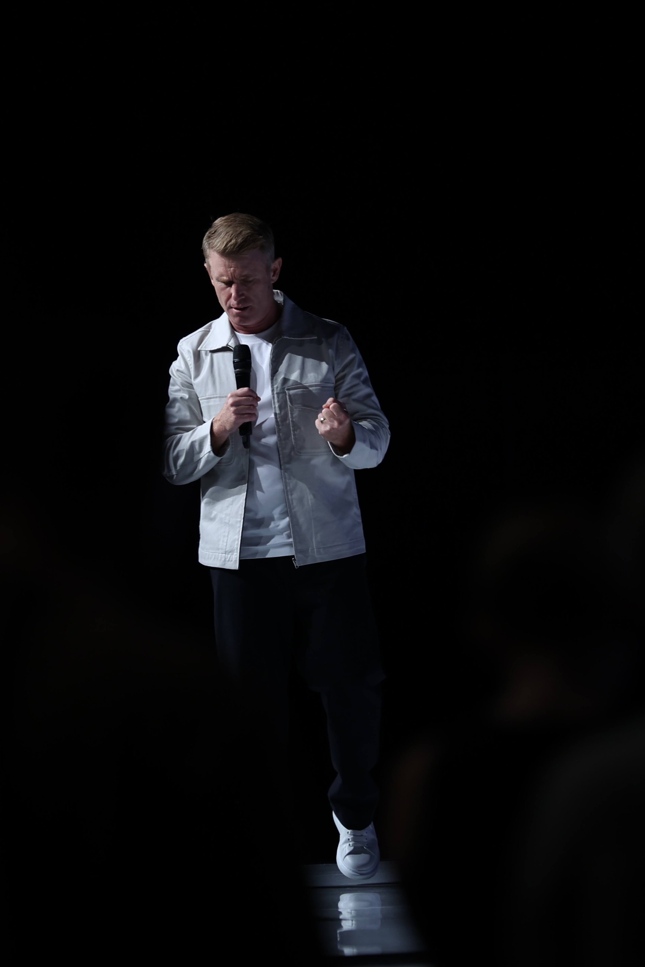 A person holding a microphone, wearing a light jacket, dark pants, and white shoes, standing against a black background.