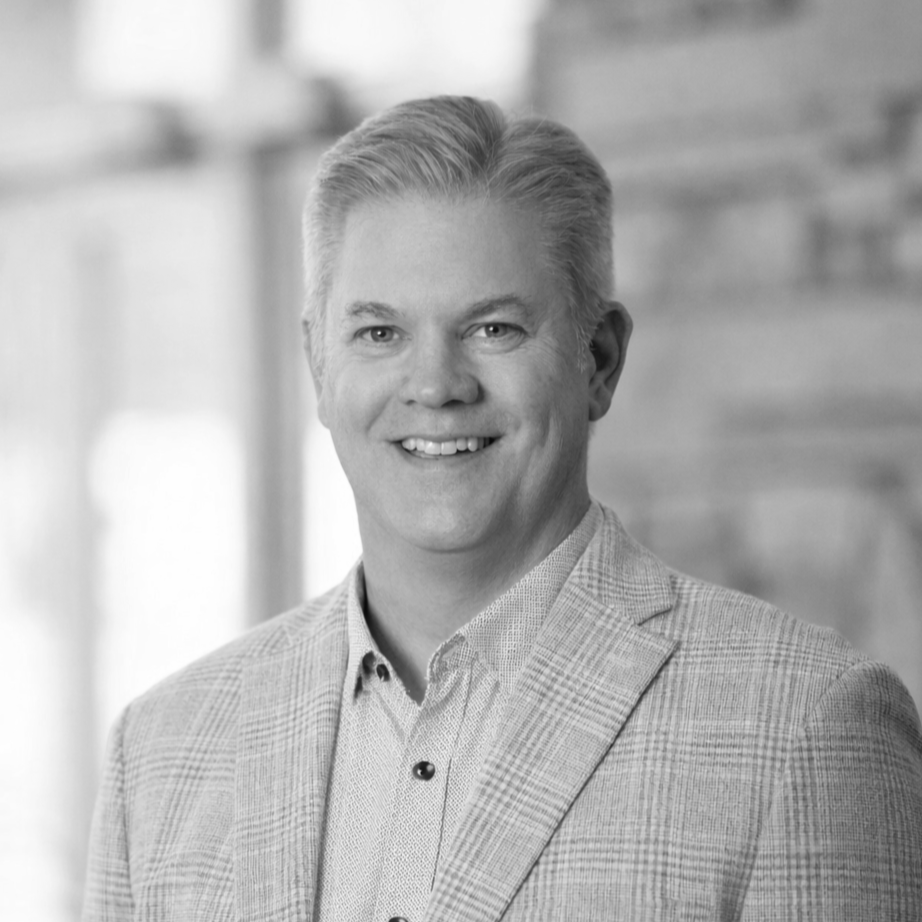 Black and white portrait of a smiling man with neatly combed hair, wearing a light-colored blazer over a collared shirt, standing in front of a blurred indoor background.