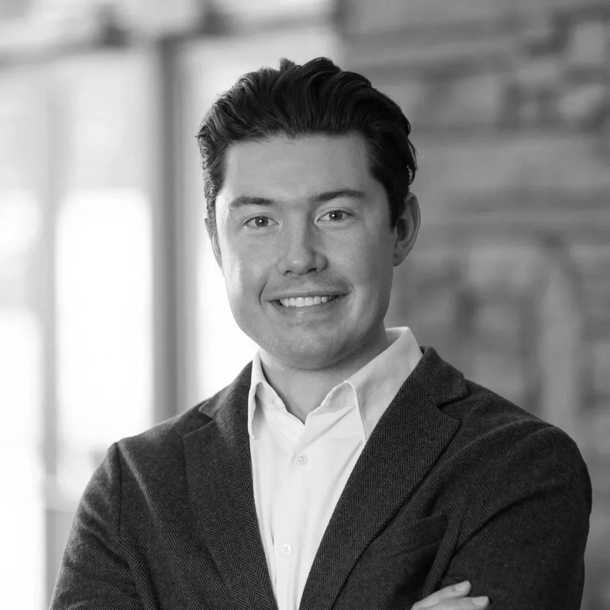 Black and white portrait of a young man with dark hair, wearing a suit jacket and white shirt, smiling and crossing his arms indoors.