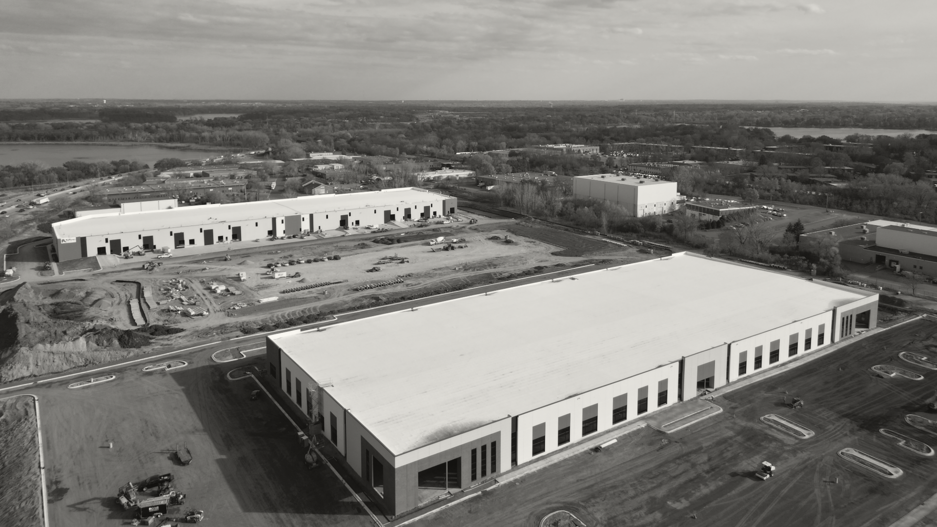 Aerial view of a construction site with multiple large warehouse buildings, some unfinished, with construction vehicles and materials scattered around. The site is surrounded by roads and other industrial buildings.