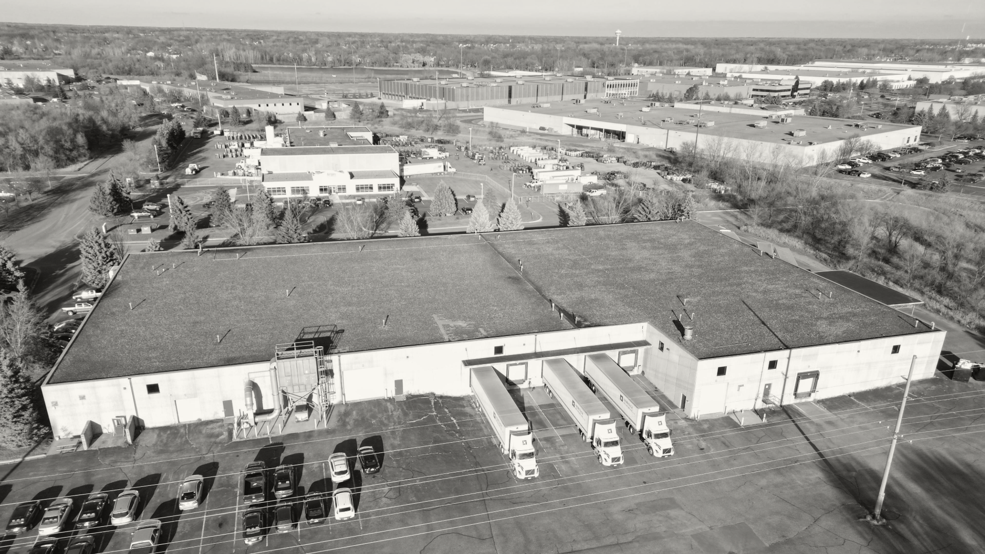 Black and white aerial view of a large warehouse building with trucks parked outside and smaller office buildings in the background. Cetra Partners Industrial Building