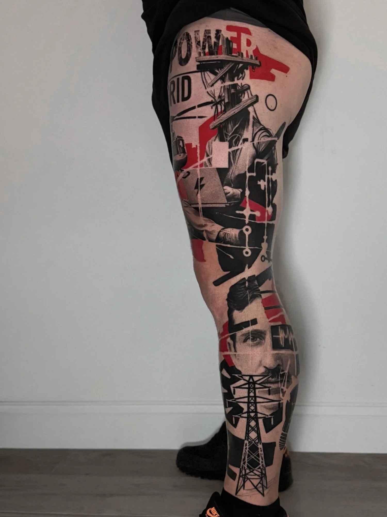 A person's leg with multiple tattoos including a portrait, a tower, and abstract designs, wearing black shoes and standing on a gray floor against a plain white wall.