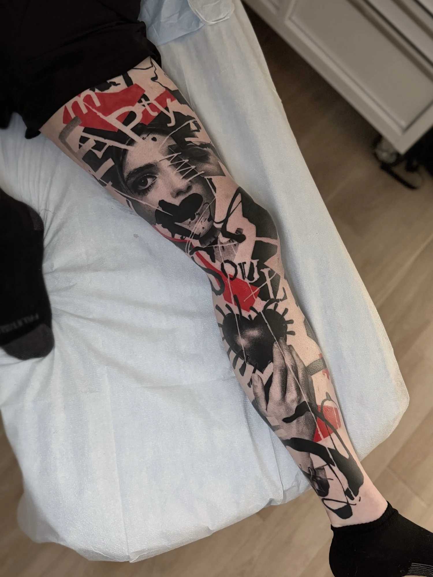 A person's leg with a colorful tattoo sleeve featuring a woman's face, abstract shapes, and red accents, lying on a white bed with a beige bedsheet.