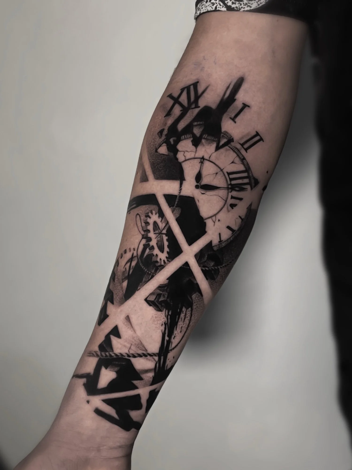 A person's forearm with a black and gray tattoo of a clock with Roman numerals and mechanical gears.