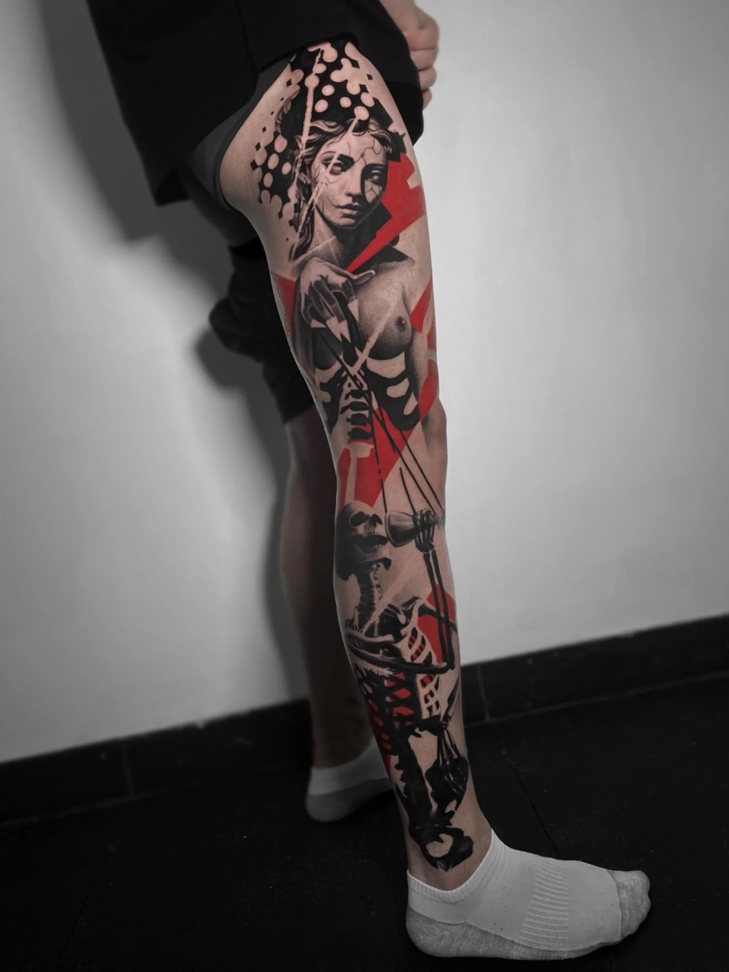 Tattooed leg with a portrait of a woman and abstract black and red shapes, standing in a room with white wall and black tiled floor.