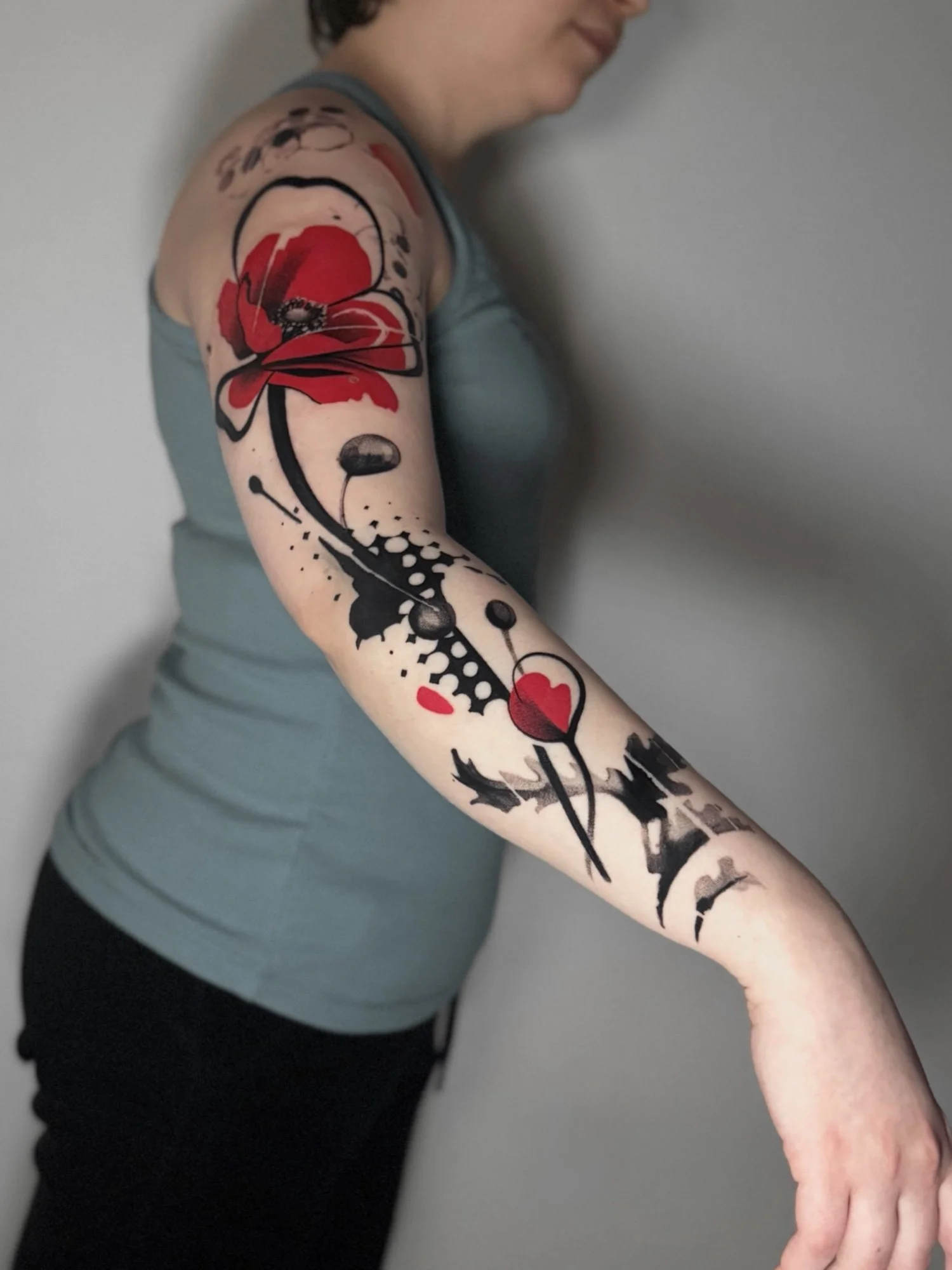 A woman with a large, colorful tattoo sleeve on her left arm. The tattoo features a red flower, black abstract shapes, and splashes.
