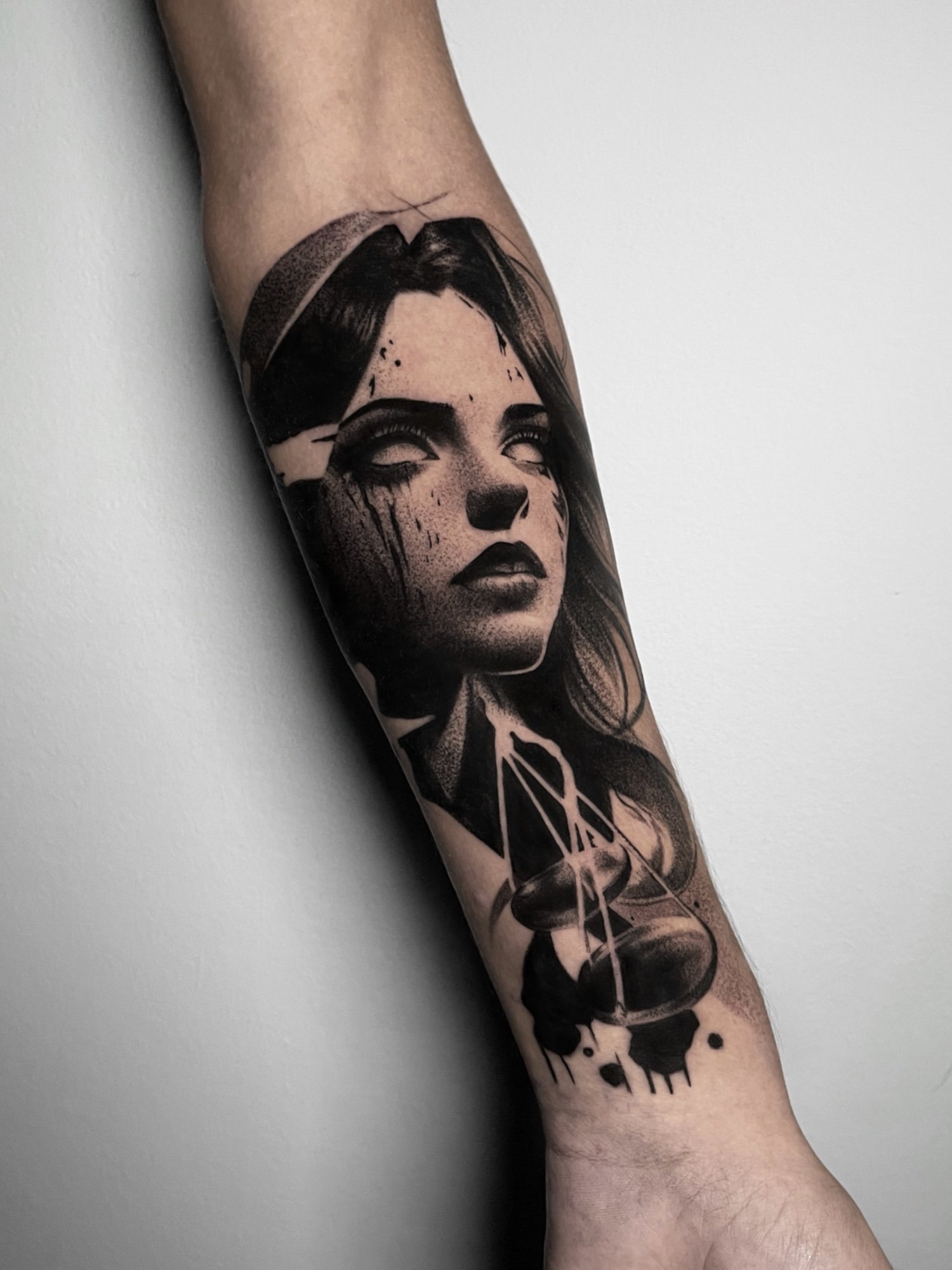 A tattoo of a woman's face on a person's forearm, with dark hair and bold makeup, combined with a stylized mushroom illustration and abstract ink splatters.