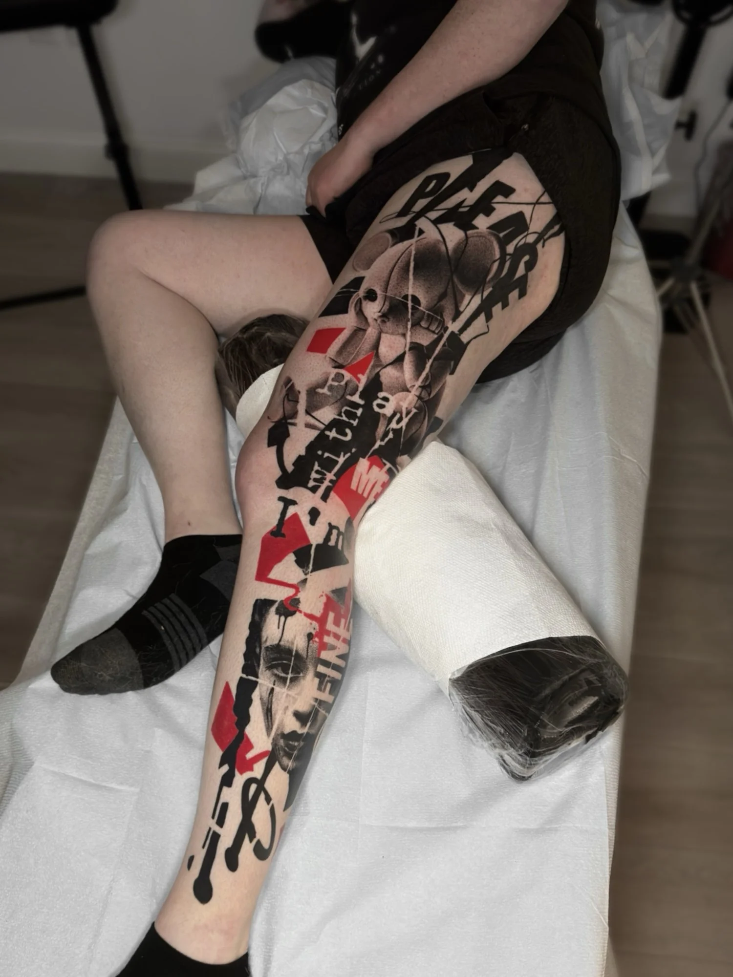 Person lying on a bed with large tattoo on their thigh and leg, wearing black shorts, black socks, and black shoes, in a clinical setting.