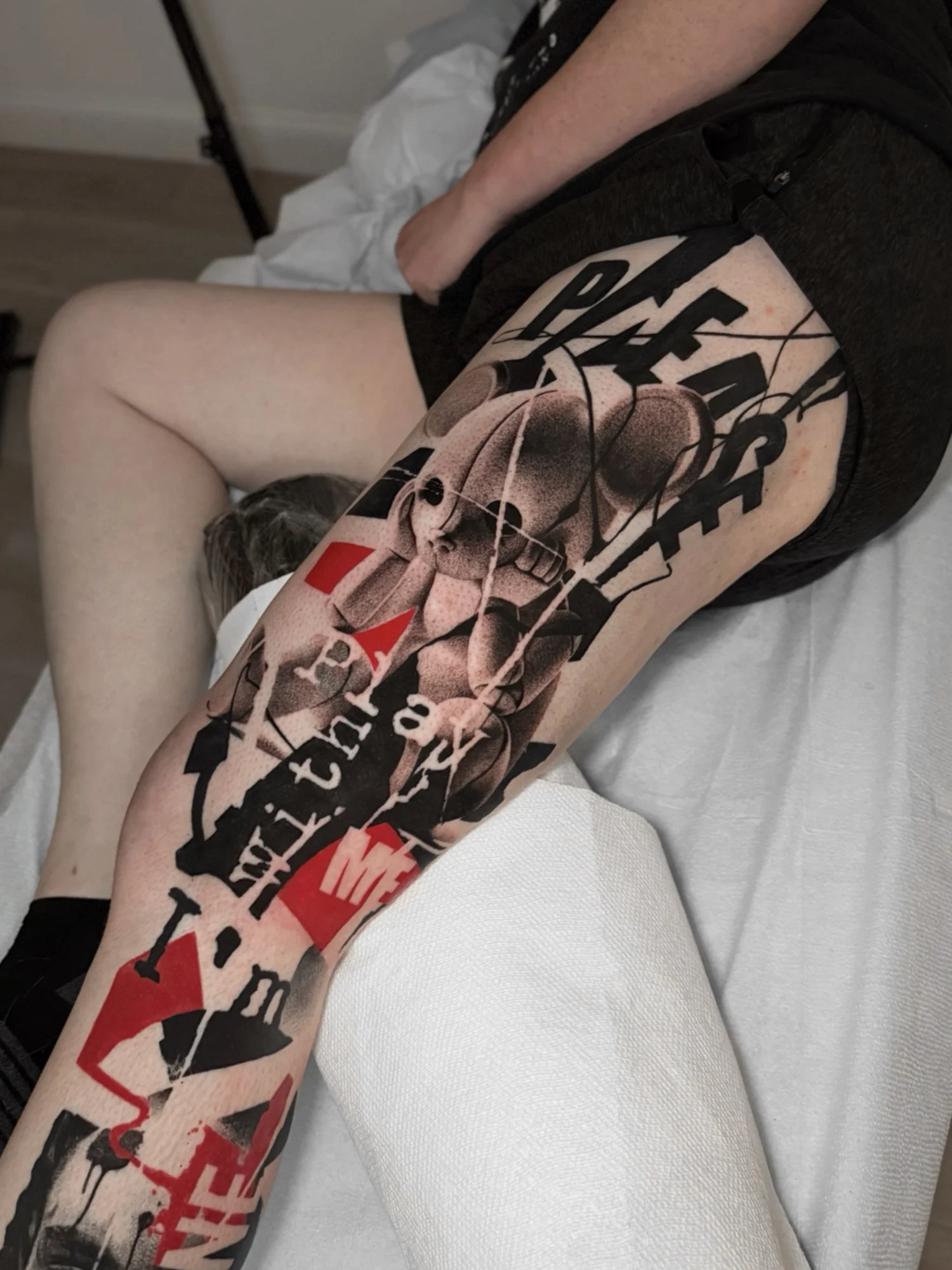 Close-up tattoo of a person's leg in trash polka style with a colorful tattoo depicting skulls, text, and abstract elements, lying on a white surface