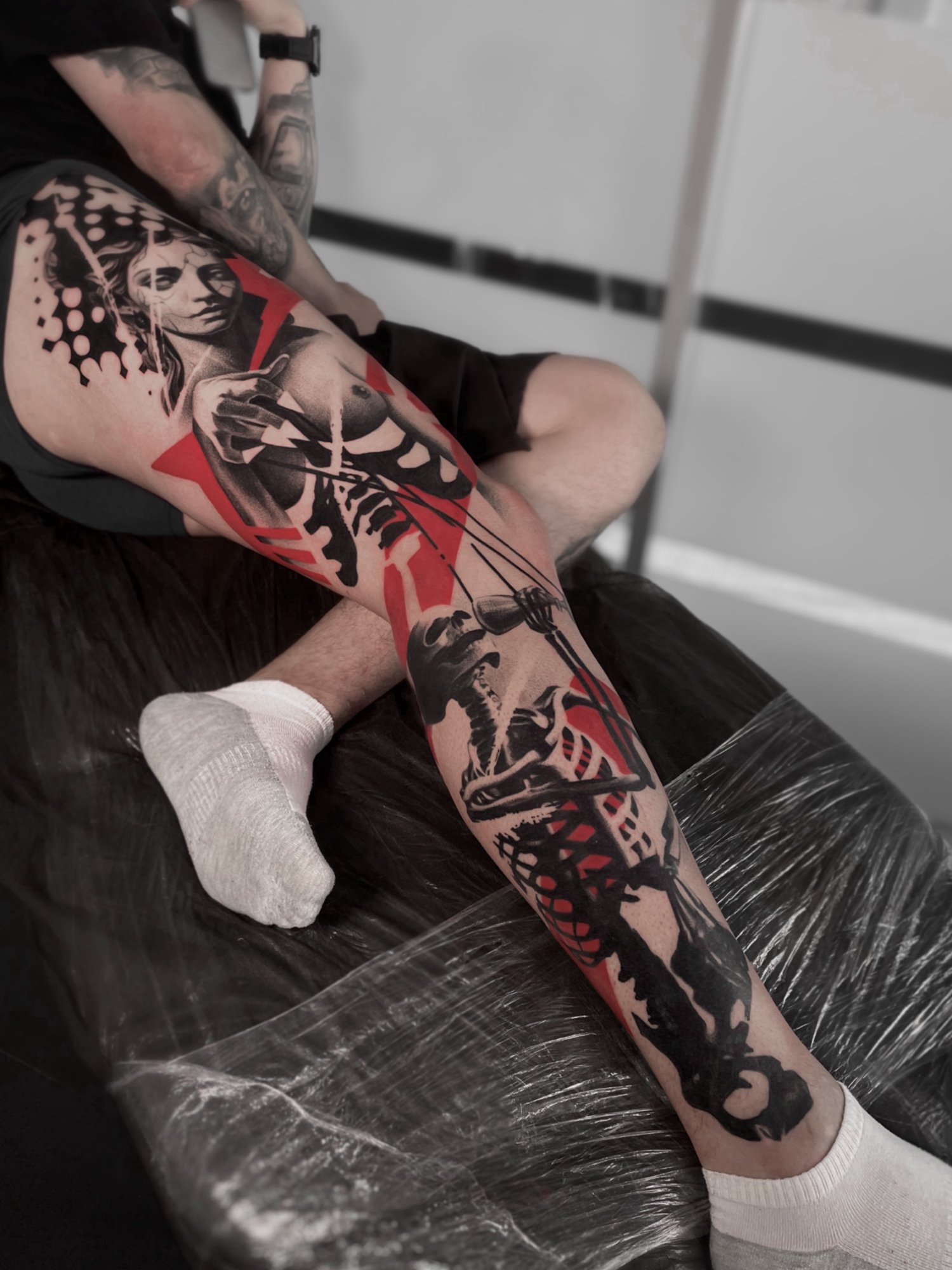 Tattooed person sitting with leg extended, displaying a large black and gray tattoo of a woman's face and abstract design on his thigh.