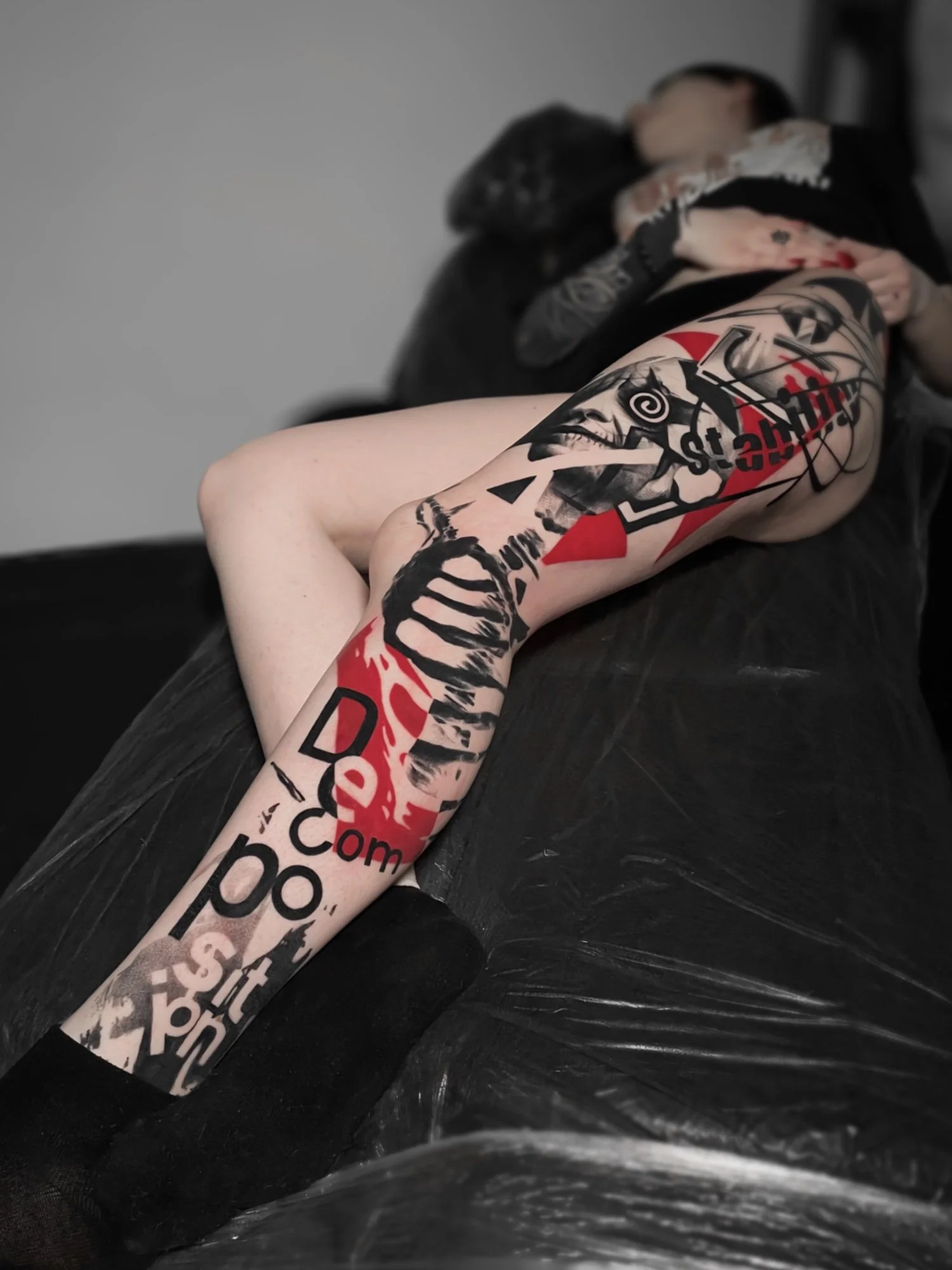 A person with a full sleeve tattoo on their left arm lying on a black surface, with the person's face partially visible in the background, resting on the person's hand.