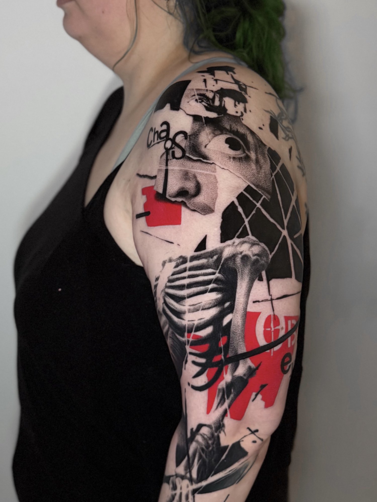 A person's shoulder and upper arm covered with a tattoo featuring a skeleton, an eye, and text, with red and black ink, against a plain background.
