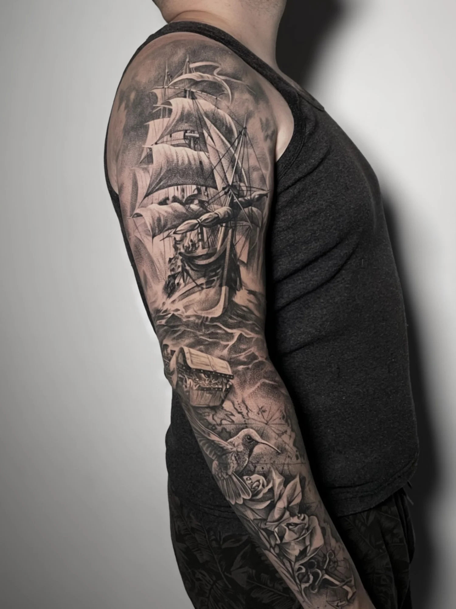 A person's arm with a detailed black and gray tattoo of a sailing ship, clouds, a boat, a bird, and roses.