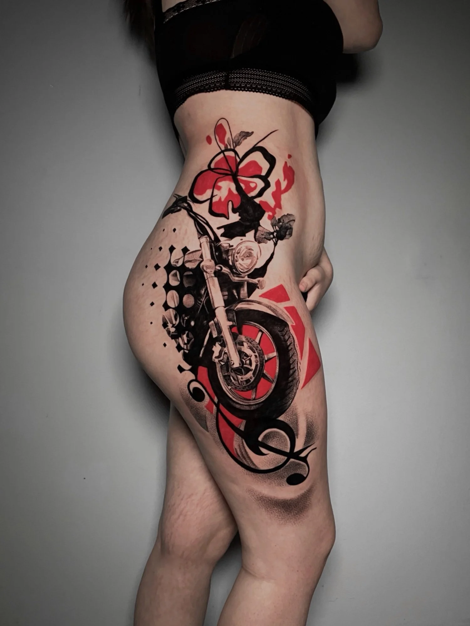 A person's thigh with a tattoo of a red and black abstract floral and motorcycle design on a gray background.