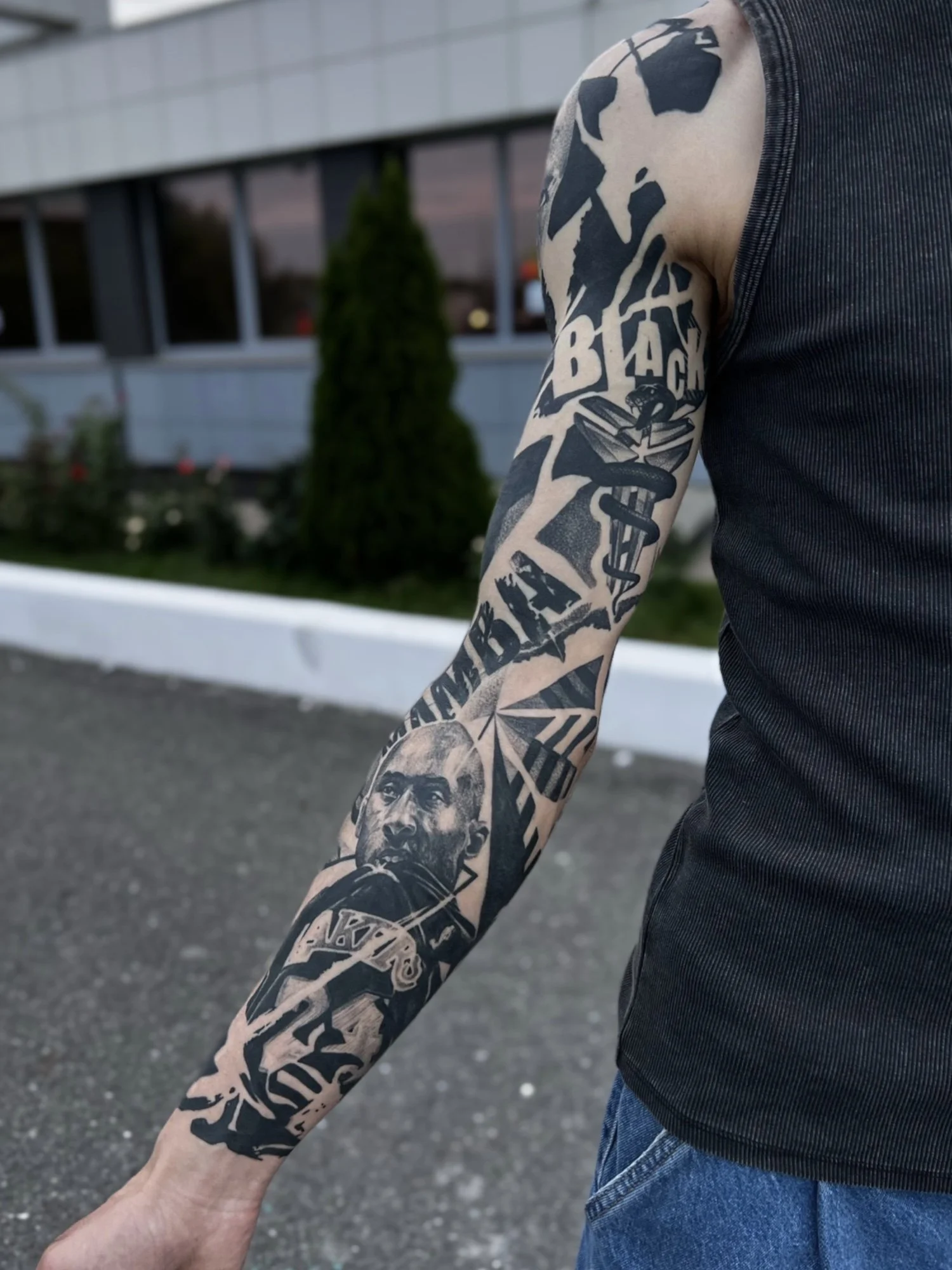 A person's arm with a large black and gray tattoo sleeve, featuring various images including a portrait of a man, text, and other designs, as they stand outdoors in front of a modern building with large windows and some greenery.