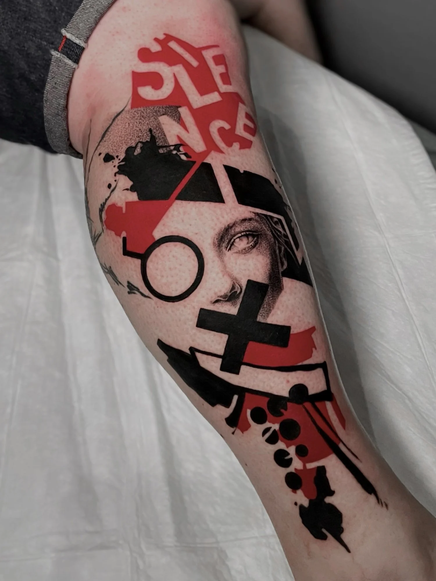 A tattoo on a person's forearm featuring a stylized face of a woman with half-closed eyes, with red, black, and grey ink abstract shapes and text around it.