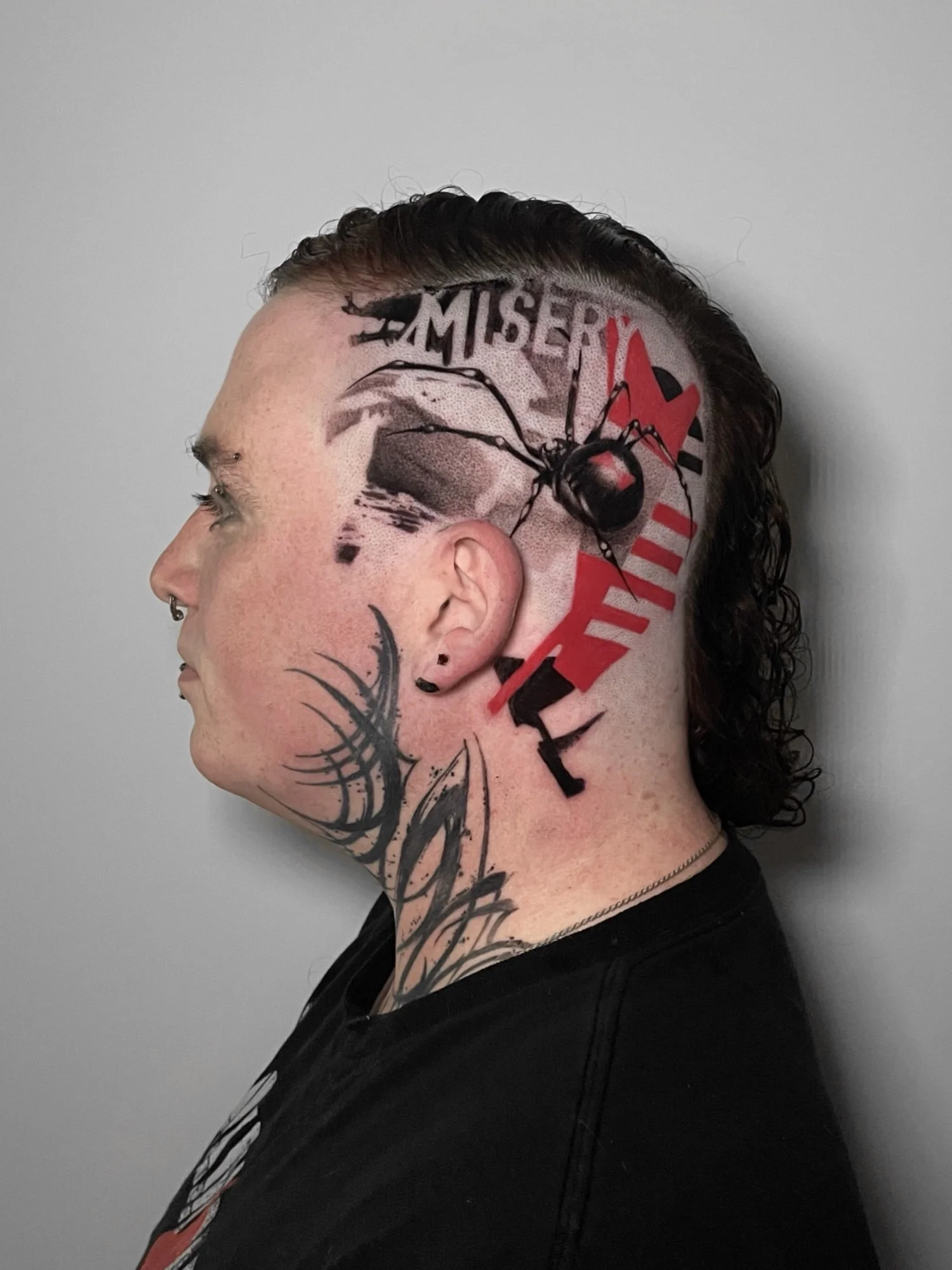 Side view of a person with a partially shaved head featuring tattoo artwork, including a large spider, the word 'MISERY,' and abstract shapes, with long curly hair on the back of the head, against a plain gray background.