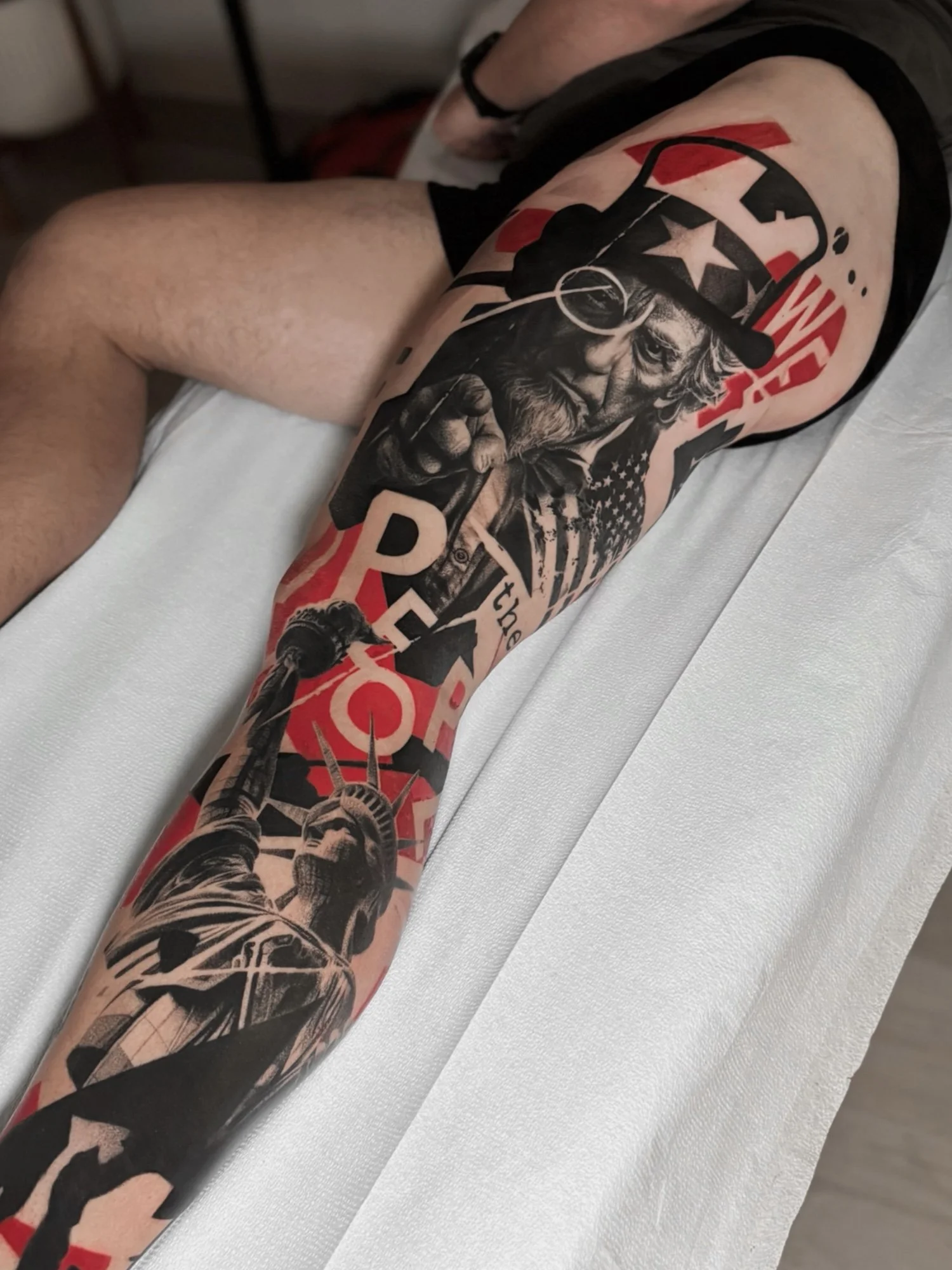 Tattooed arm featuring a detailed portrait of an elderly man with a beard and a top hat, holding what appears to be a torch, with American flag elements and various text and graphics in red, black, and white.