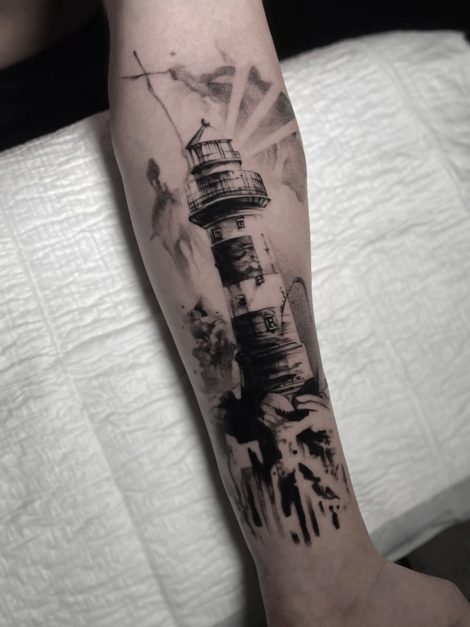 Black and gray tattoo of a lighthouse on person's forearm, with rays of light and clouds, on light skin.