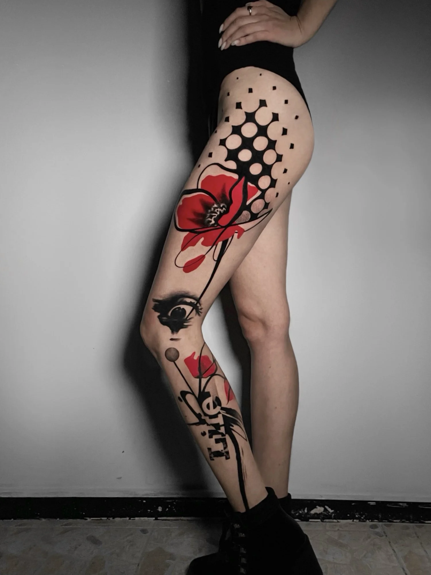 A person's tattooed leg with a large red and black flower design, black ink patterns, and abstract elements, standing against a gray wall.