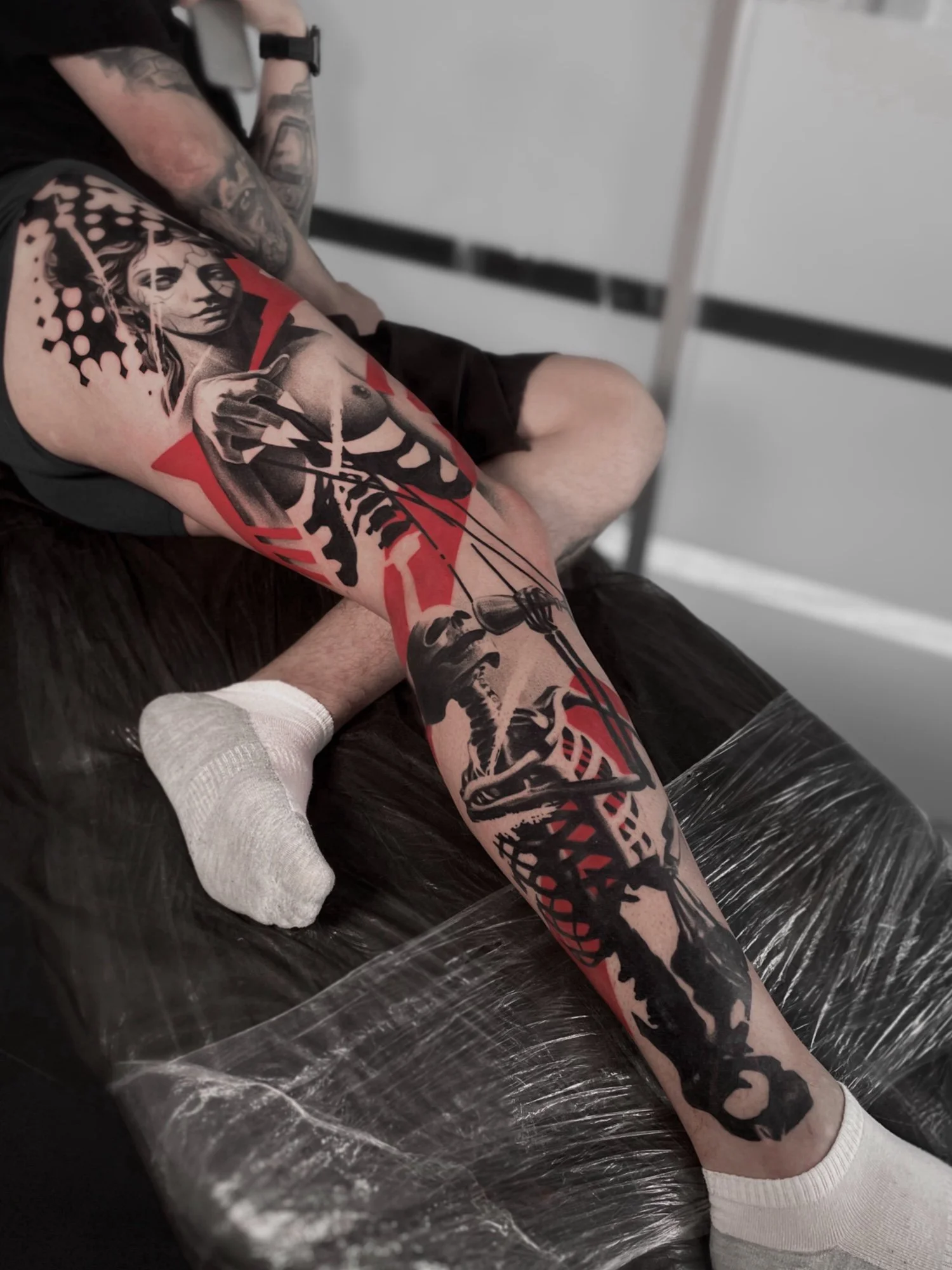 A person with tattoos on their arm is getting a black and red tattoo on their leg.