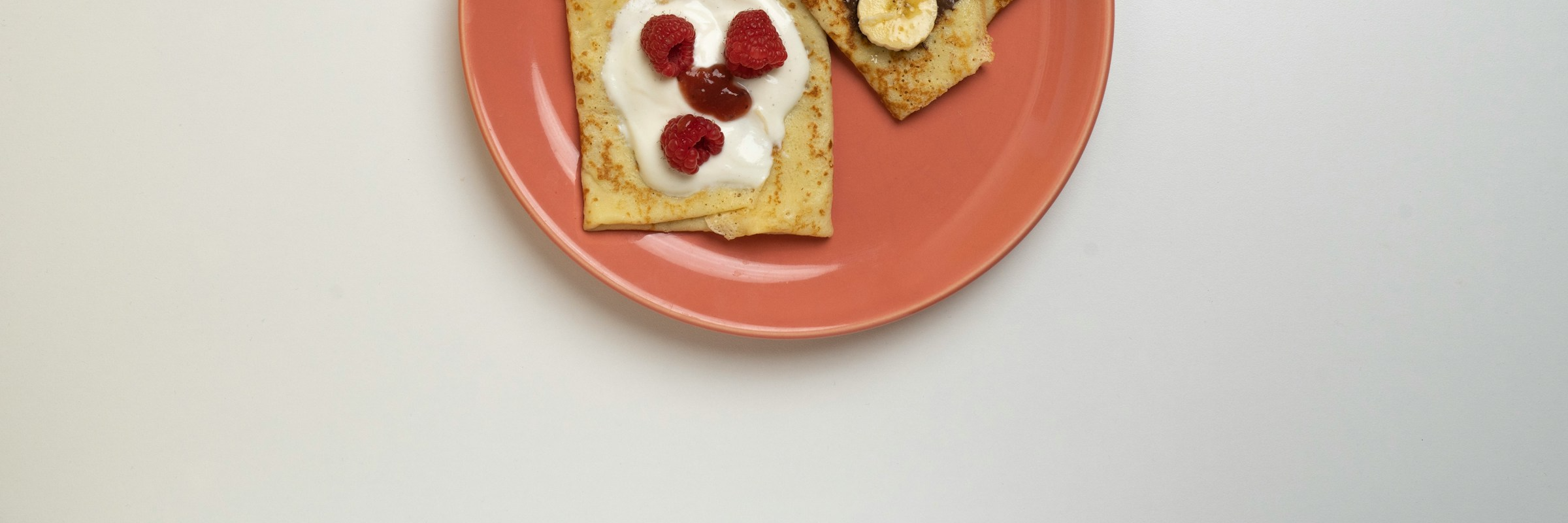 Crepes on a pink plate with raspberries, bananas, yogurt, and jam.