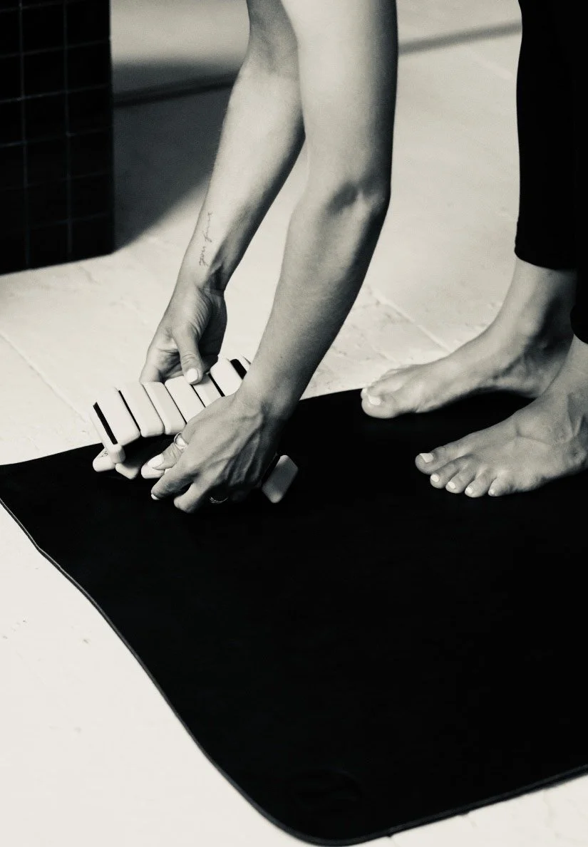 A person with a tattoo on their left forearm is bending down, placing or adjusting a small keyboard instrument on a yoga mat. The person's toes are visible and the background includes a tiled surface.