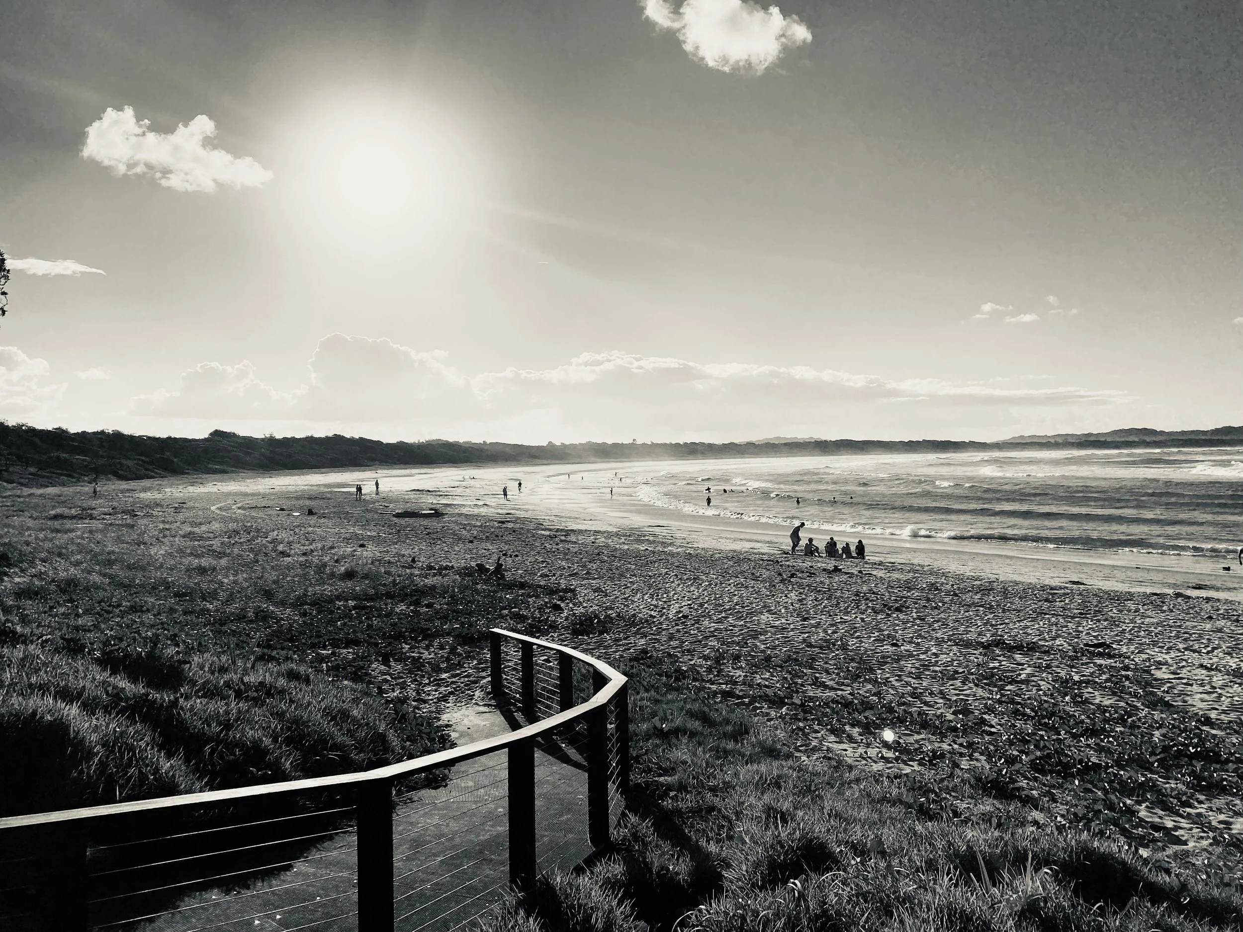 Black and white photo of a beach with people near the shoreline, a wooden fence in the foreground, and the sun shining brightly in the sky with some clouds.