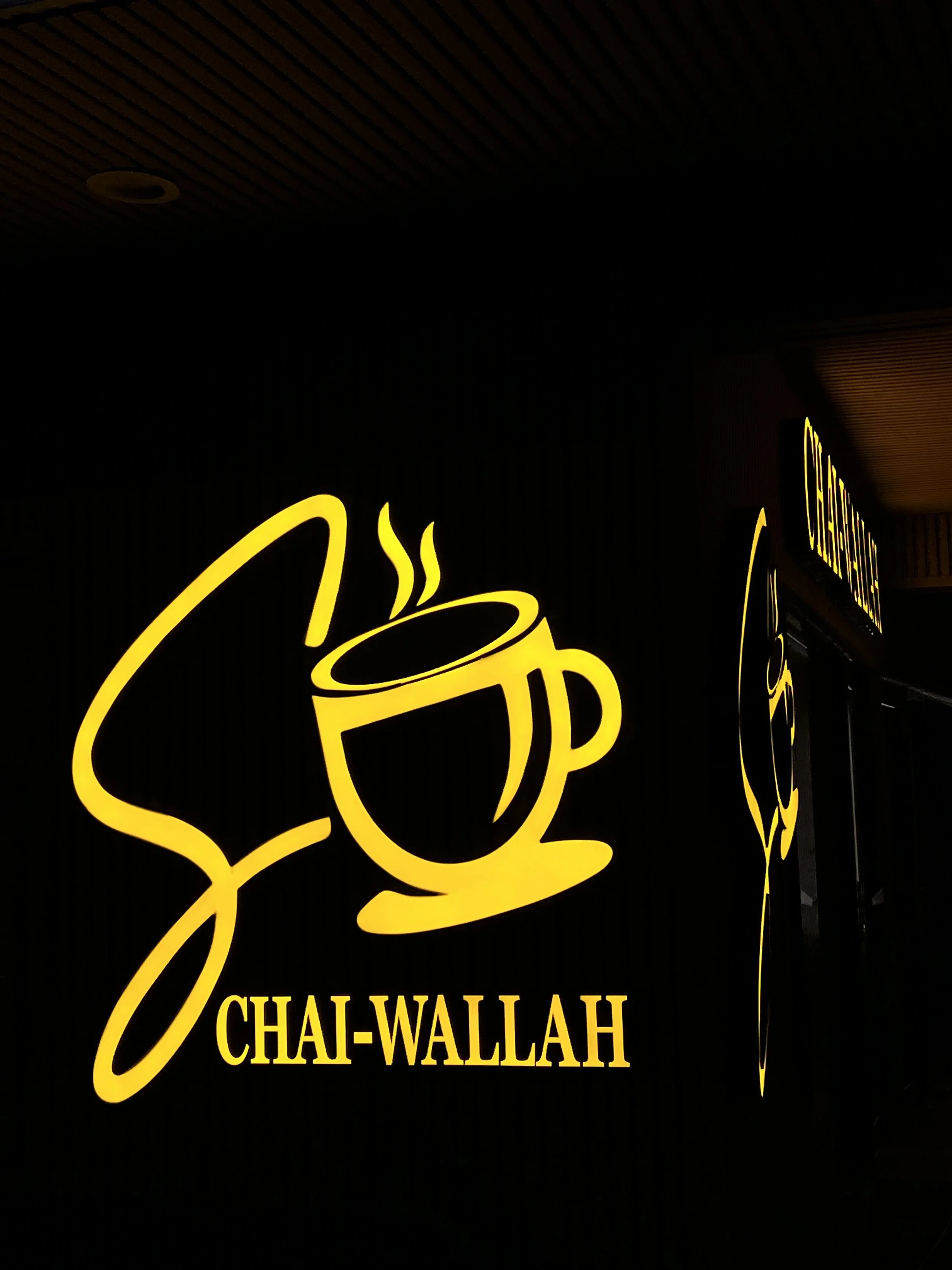 Yellow illuminated sign featuring a coffee cup with steam and the words "Chai-Wallah" underneath.