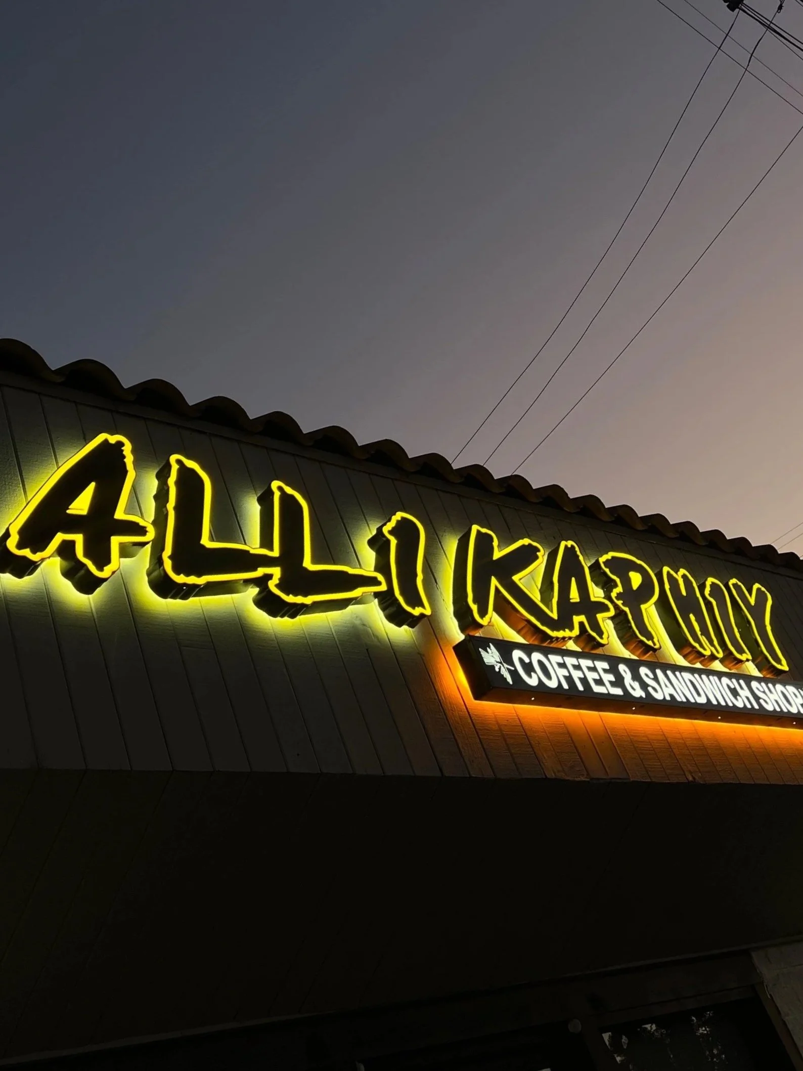 Neon sign for Ally Kapo, a coffee and sandwich shop, against a dusky sky with power lines in the background.