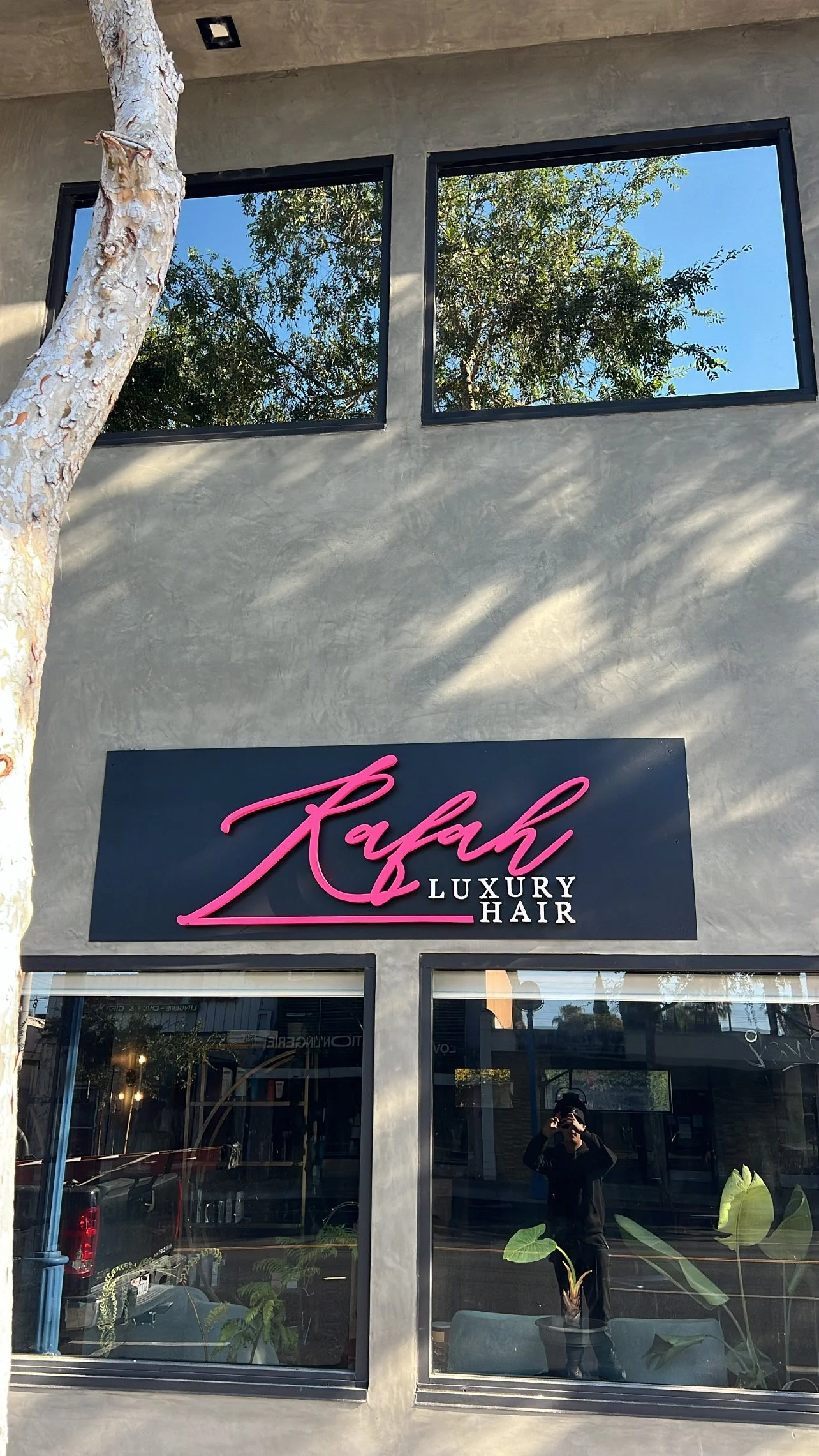Storefront sign reading 'Rafah Luxury Hair' in black and pink lettering, with two upper windows reflecting trees and a blue sky, and two lower glass windows showing the interior and the person taking the photo.