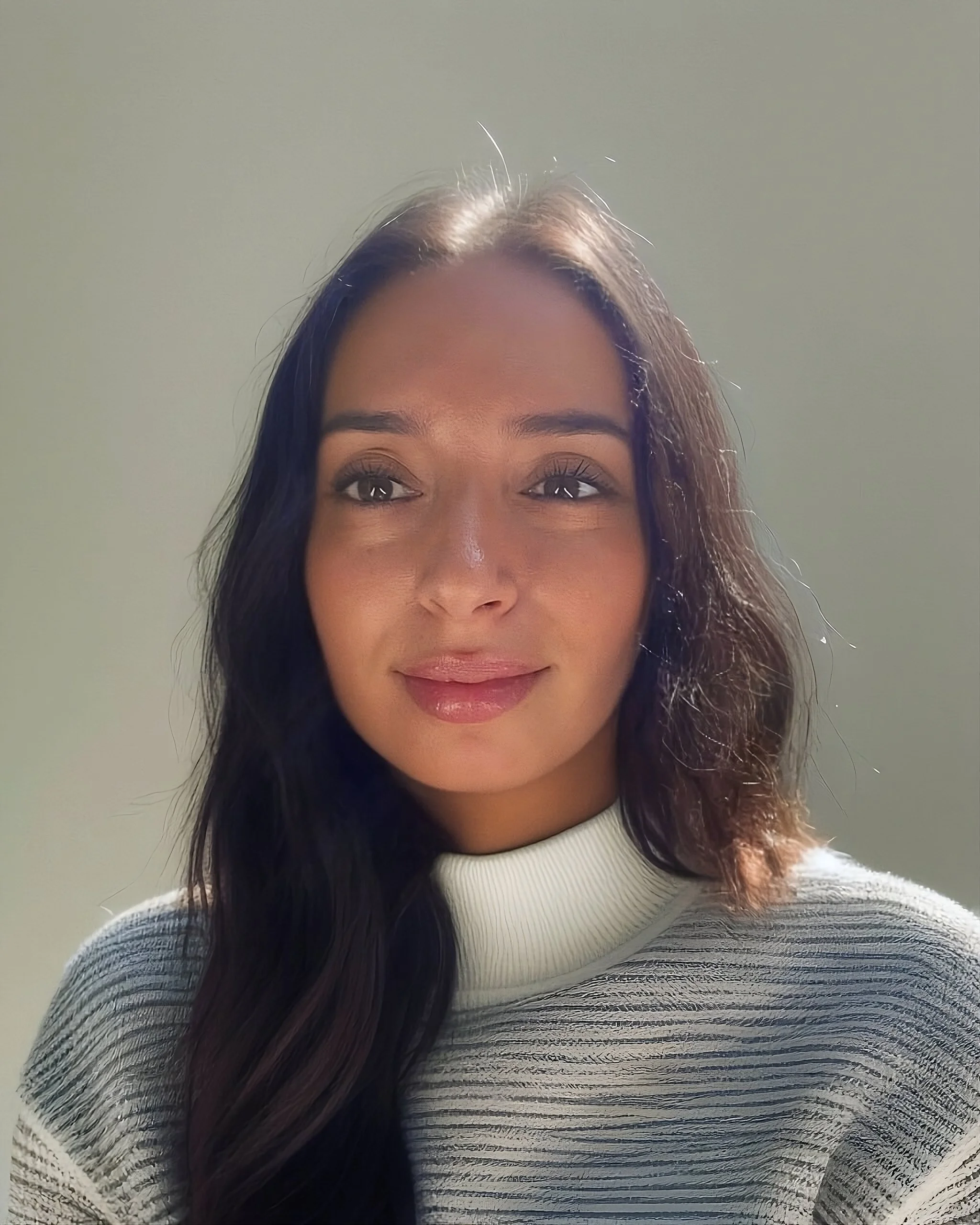 A woman with long, wavy dark hair, wearing a white ribbed turtleneck sweater, smiling softly, against a plain light-colored background.