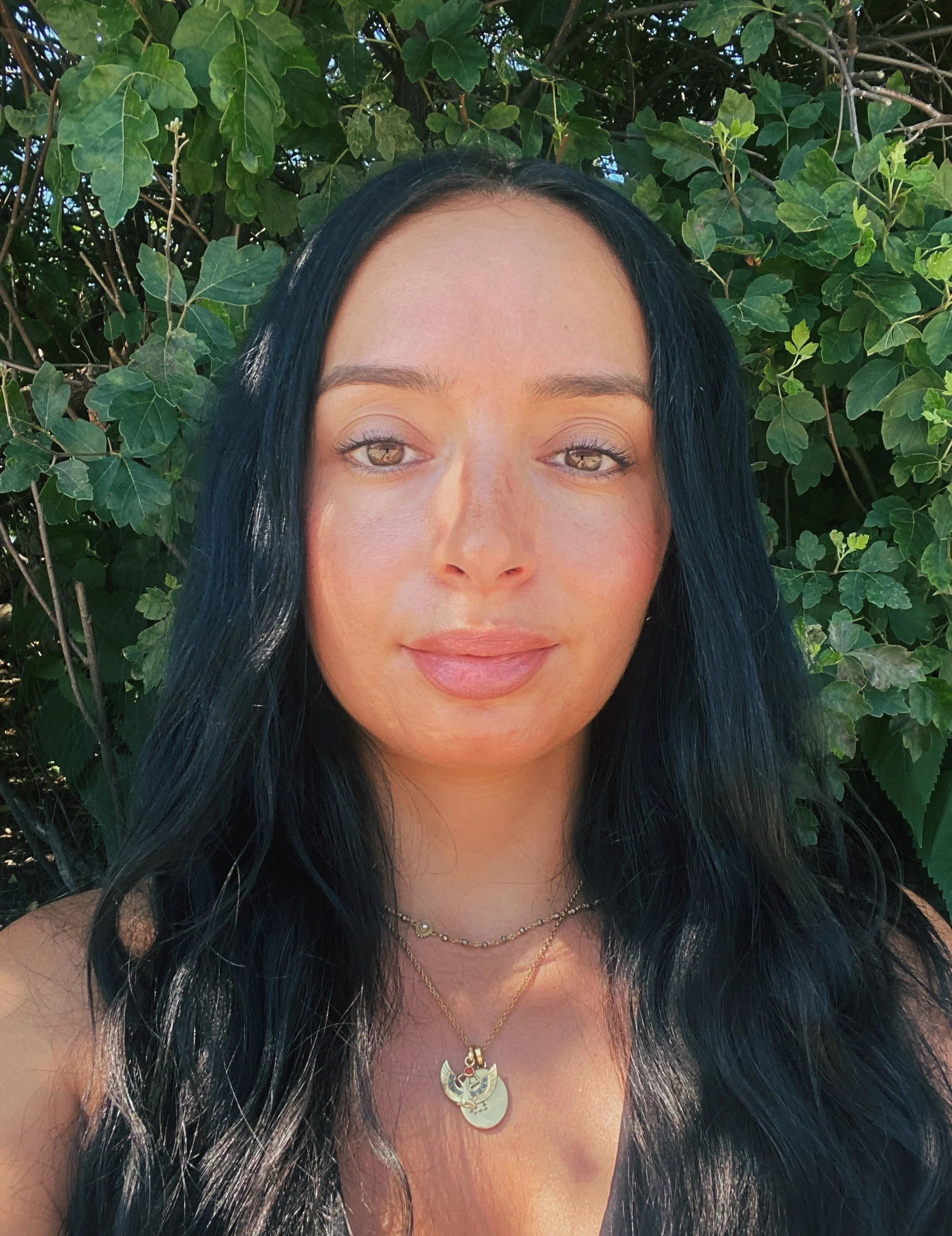 A woman with long black hair and light makeup, standing outdoors in front of green leafy plants.
