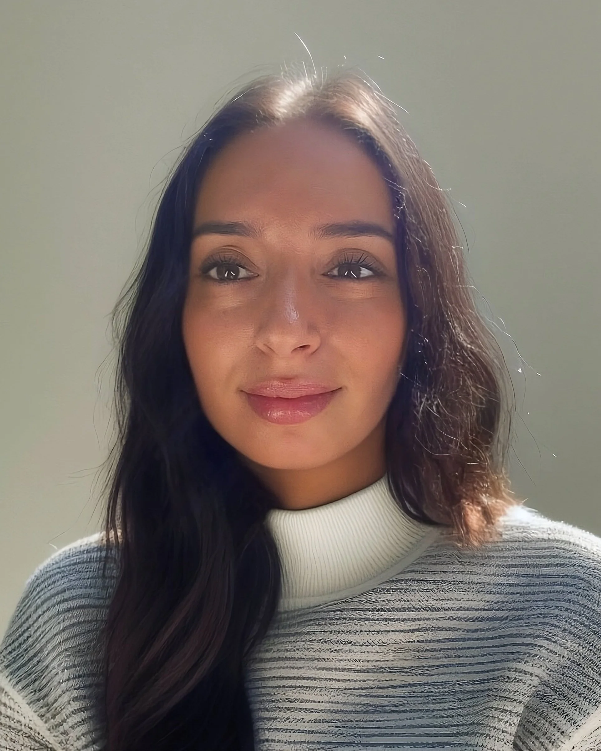 Close-up of a woman with long wavy dark hair wearing a white and gray striped sweater, looking slightly to the side with a soft smile, illuminated by natural light.