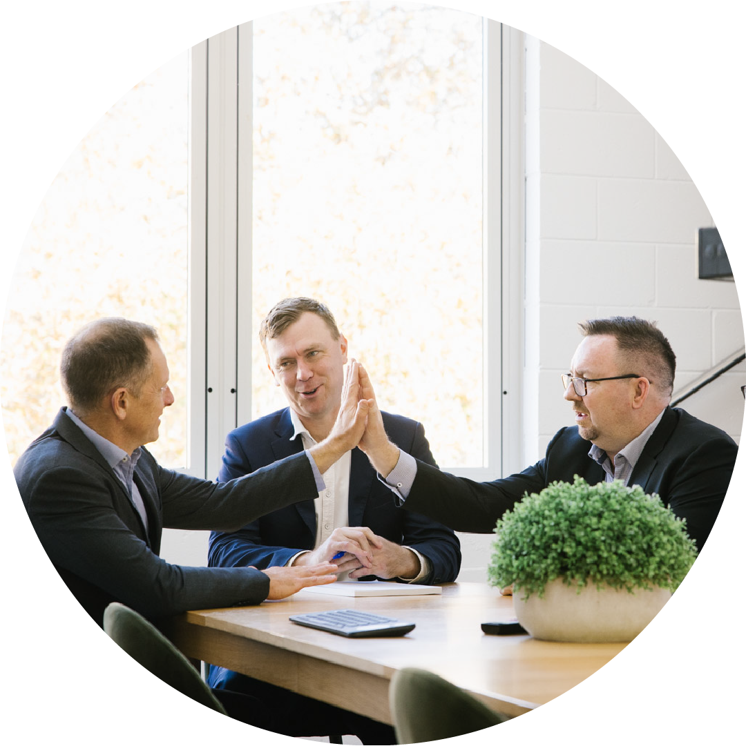 Three men in business suits sitting at a wooden table in a conference room, engaged in a friendly high-five, with a window showing trees in the background and a potted plant on the table.