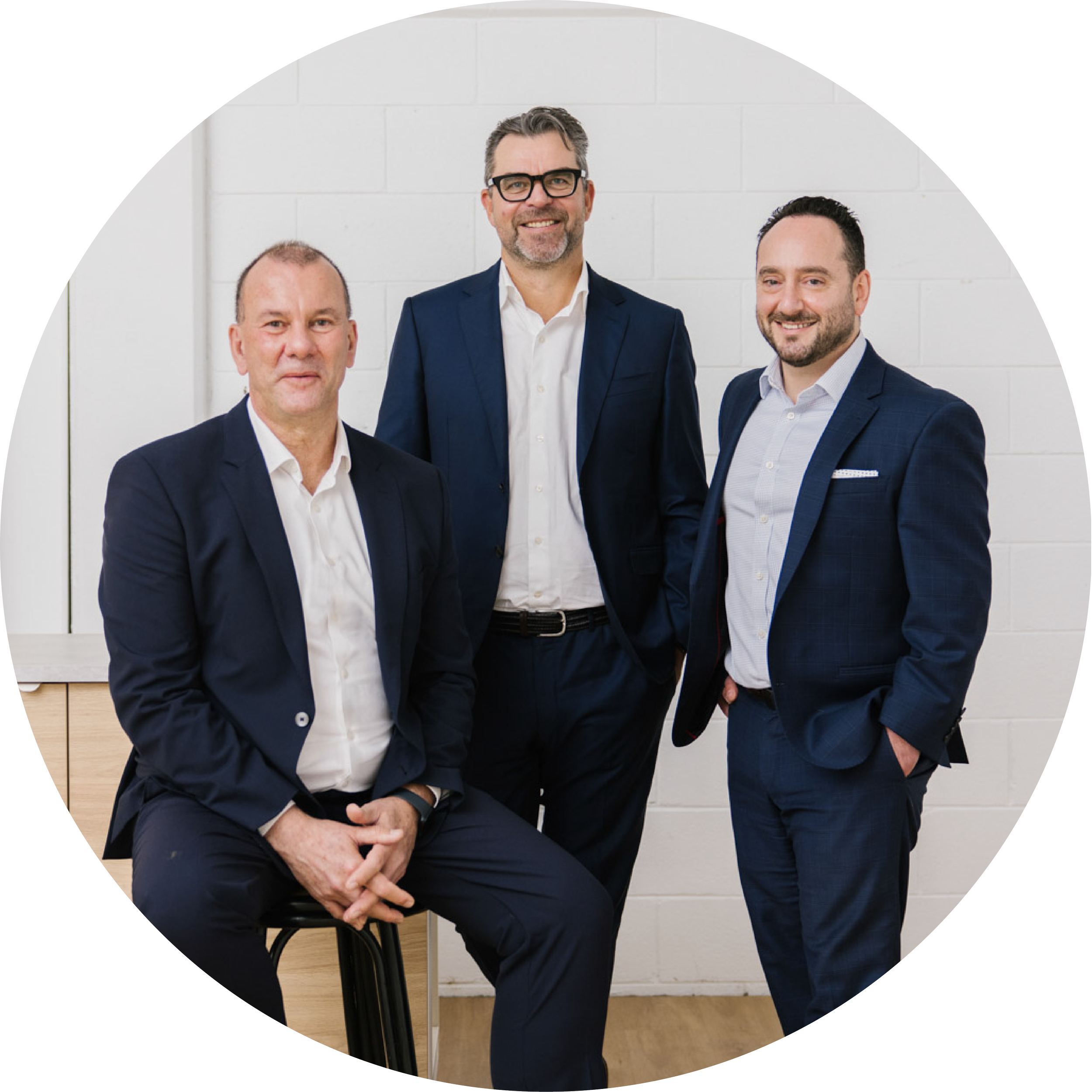 Three men in business suits standing and sitting in front of a white wall, smiling for a professional photo.