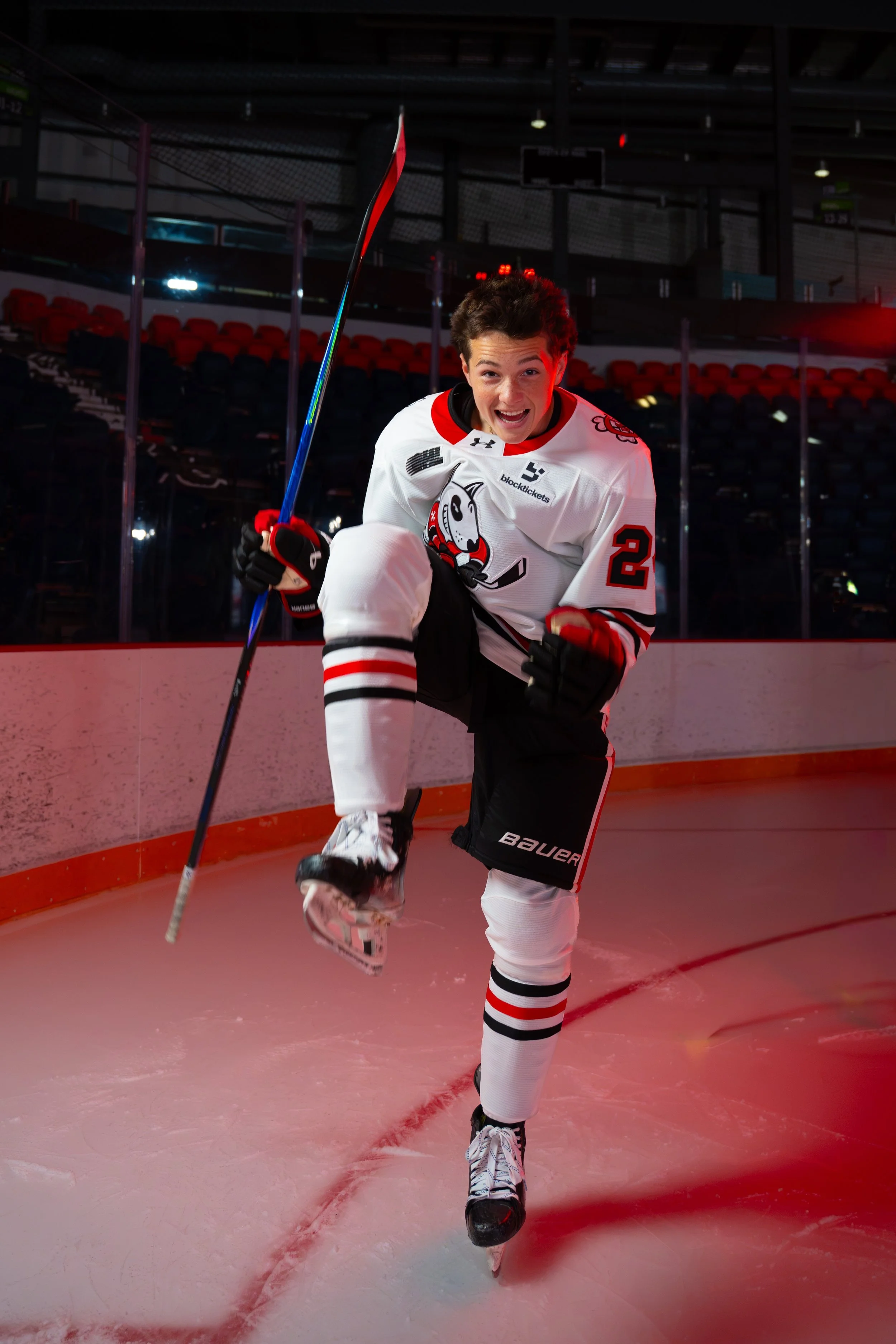 Young male hockey player in a white jersey with black and red accents, smiling and celebrating on the ice rink, wearing hockey gear including skates and gloves, with a hockey stick raised in one hand.