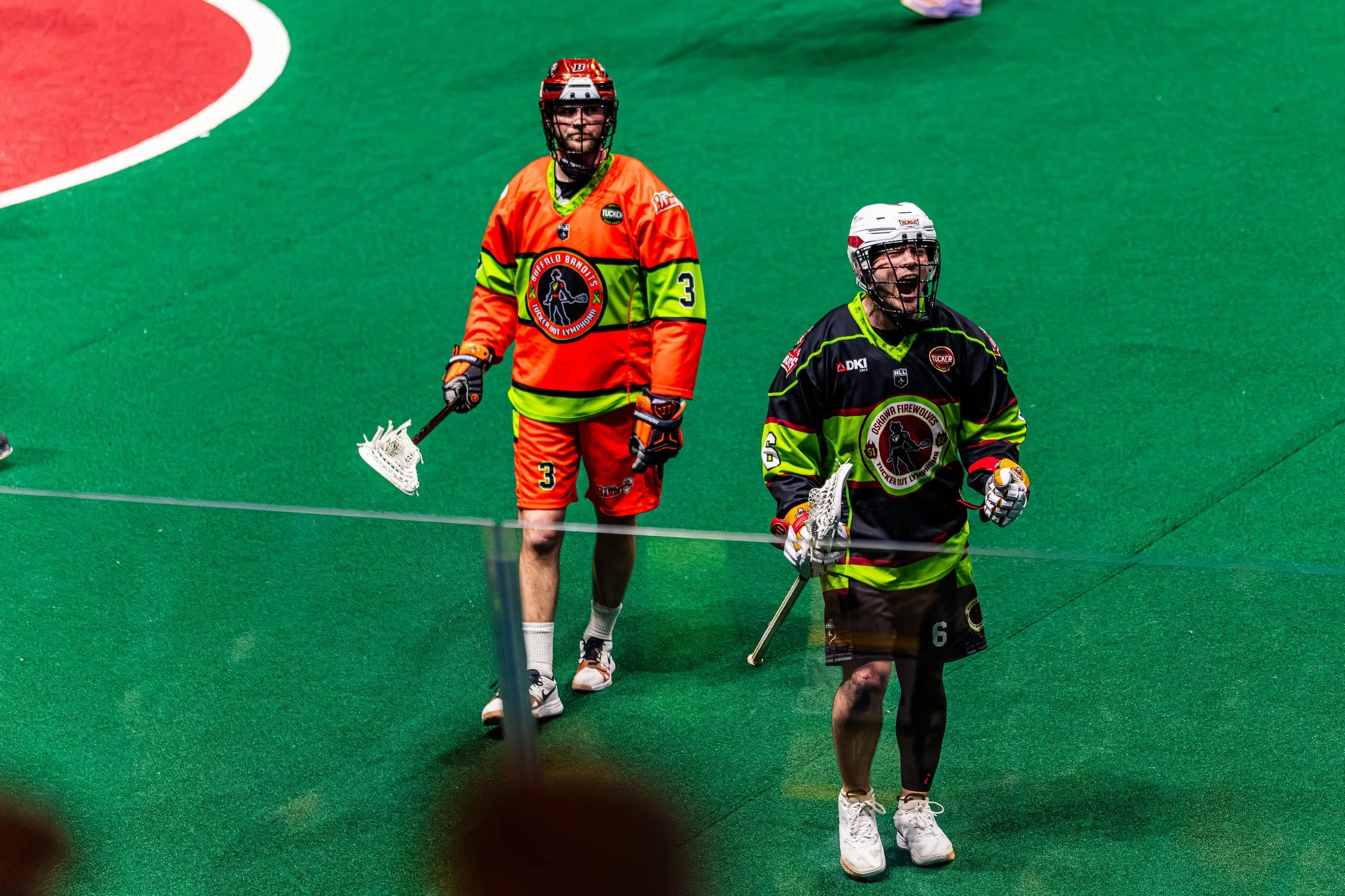 Two lacrosse players in colorful jerseys and helmets on a green turf field, one with mouth open, holding lacrosse sticks.