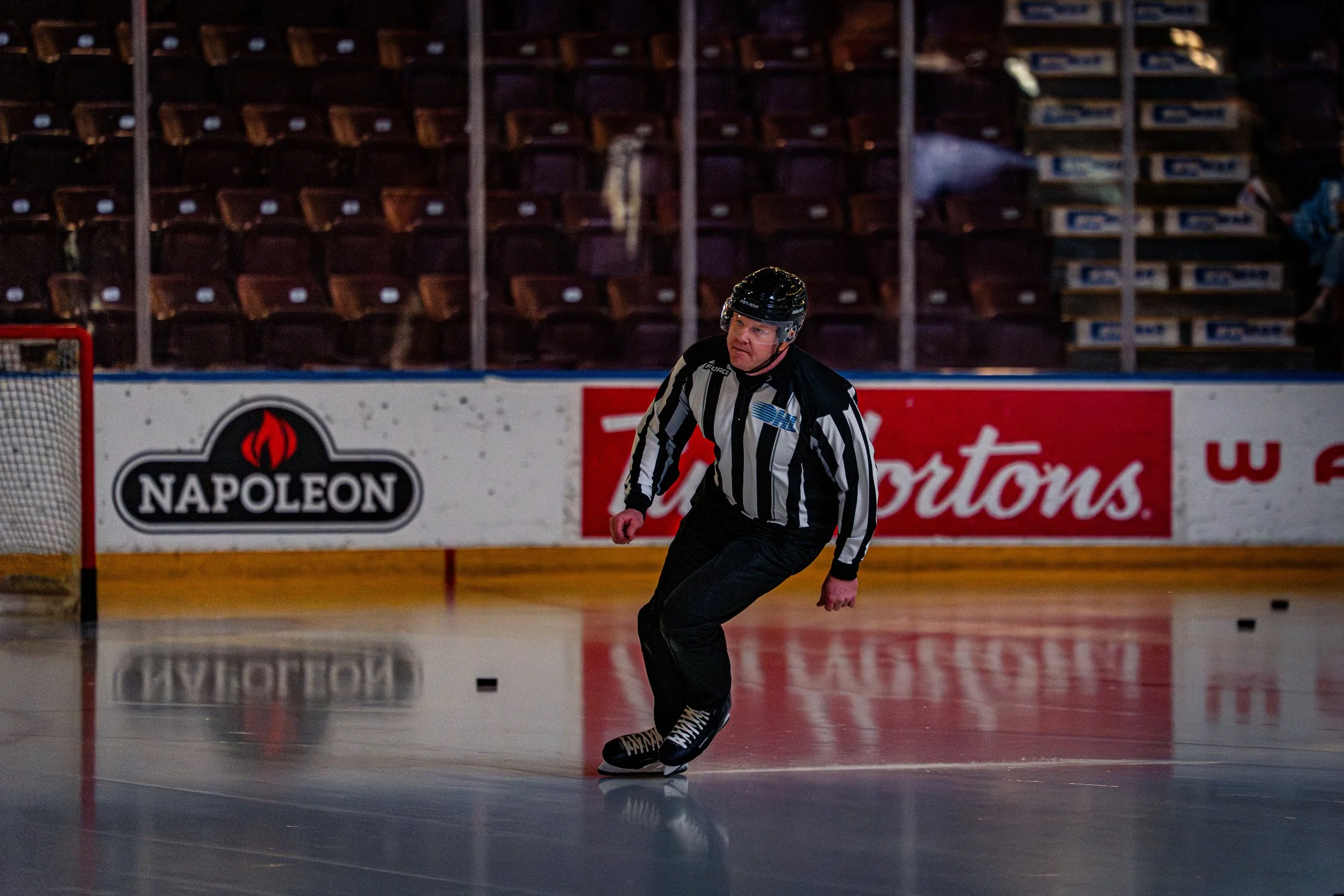 A hockey referee skating on the ice rink during a game, with empty seats and advertisements in the background.